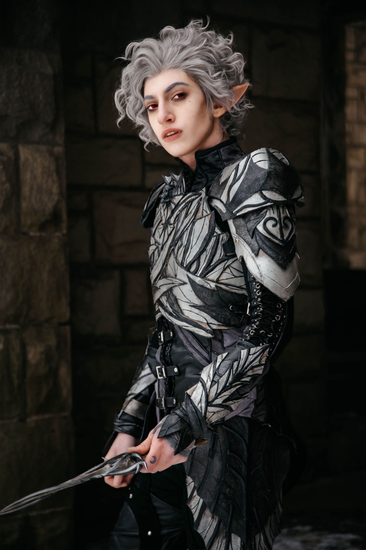 Person cosplaying as a dark elf warrior with gray curly hair, pointed ears, black armor, and a dagger.