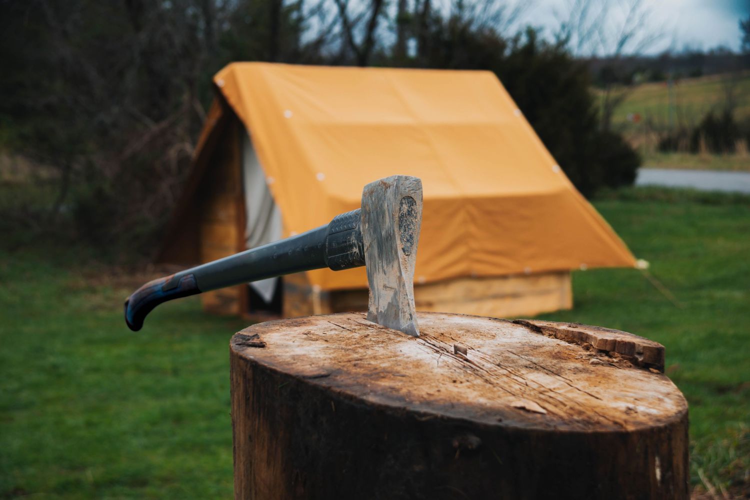 An axe embedded in a wooden stump at a campsite with a yellow tent in the background.
