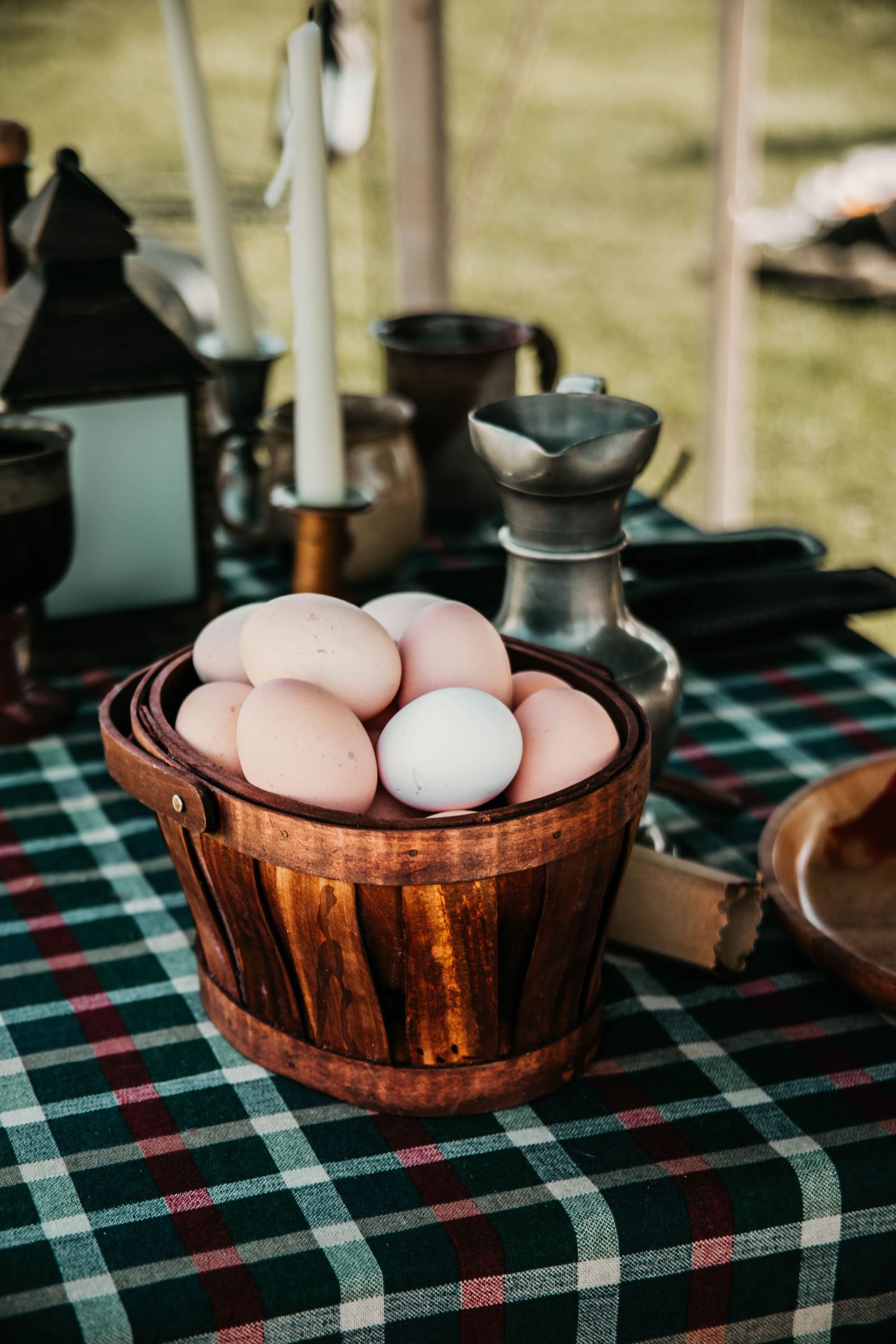 White eggs rest in a wooden bowl on a plaid tablecloth in a rustic outdoor setting.