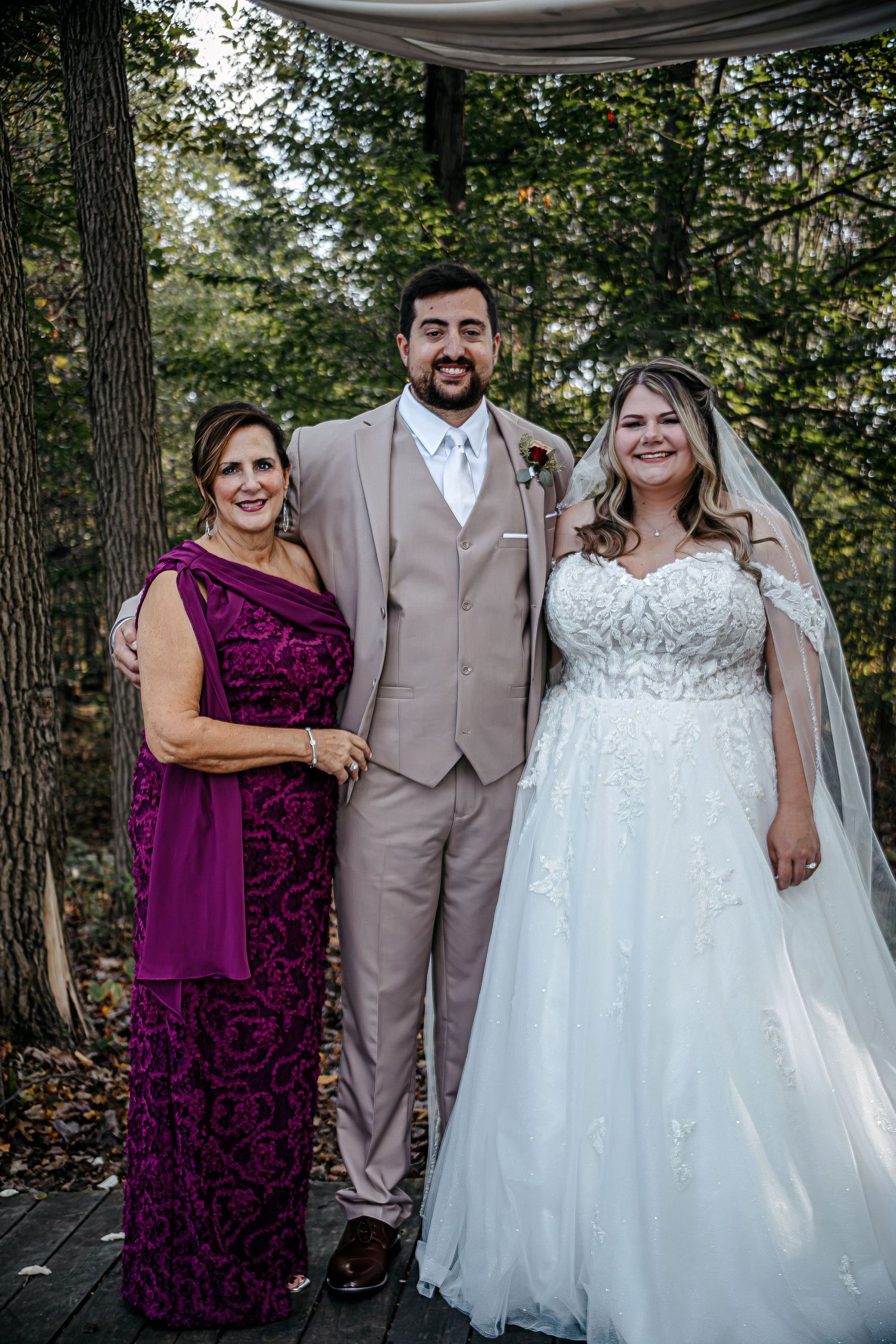 Wedding party members in purple dresses and tan suits pose under a draped canopy outdoors.