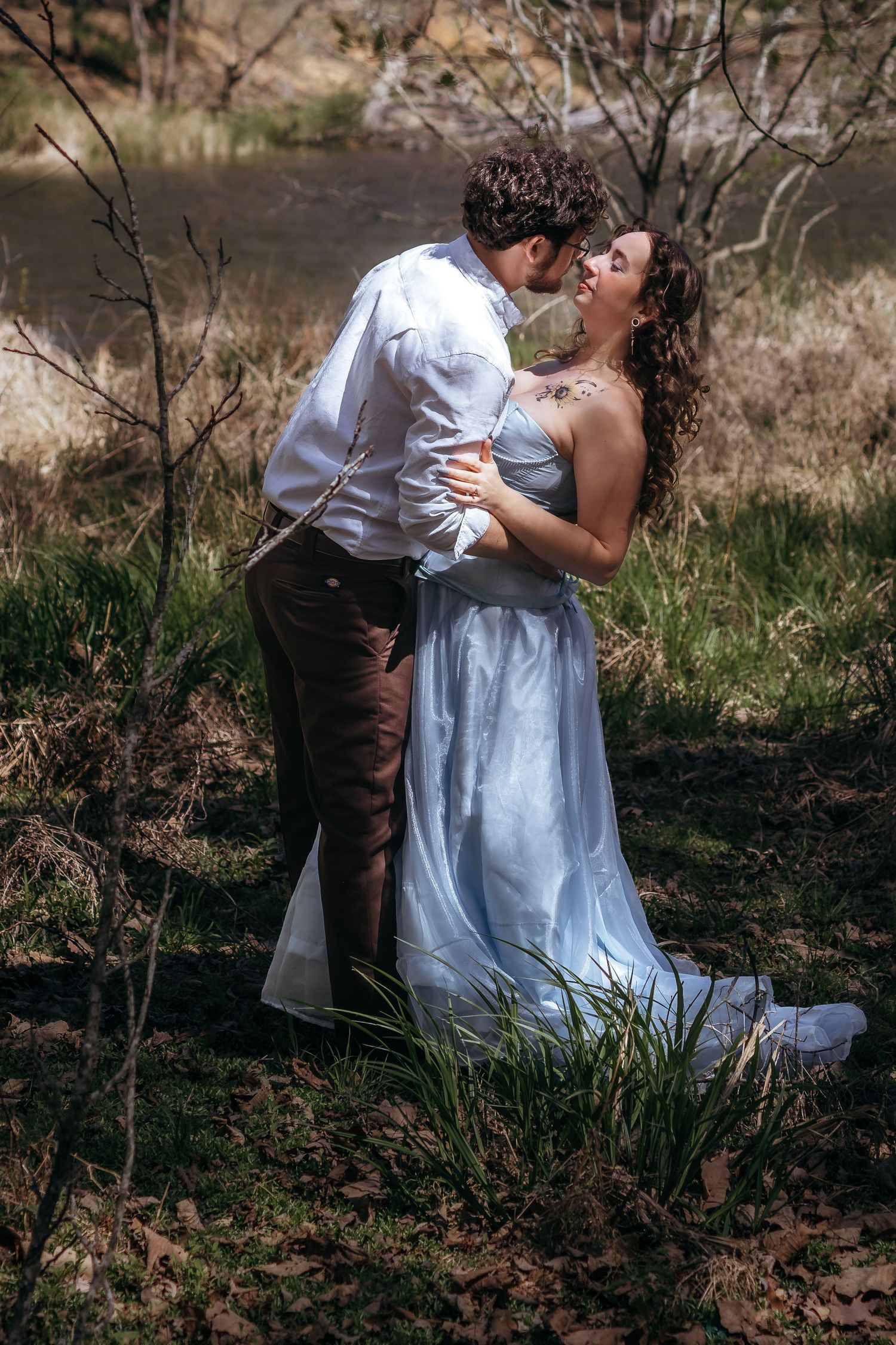 A couple shares a romantic kiss in a woodland setting, the bride in a flowing light blue gown embracing her groom.