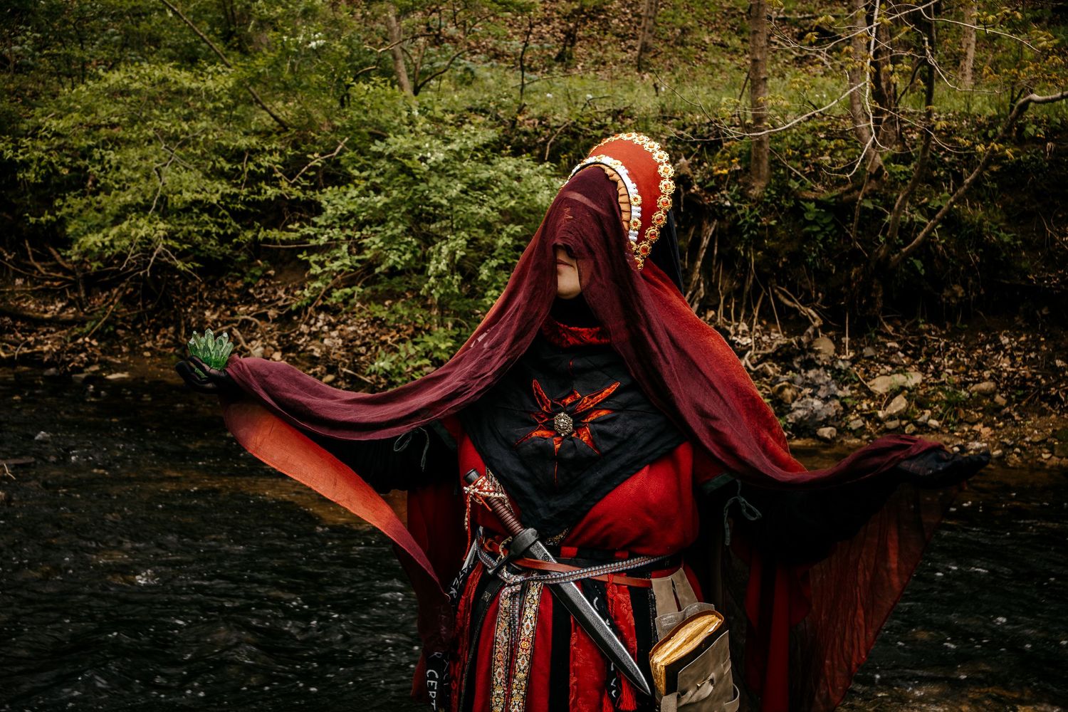 Person in red and black fantasy cosplay costume with hooded cloak and sword poses by a forest stream.