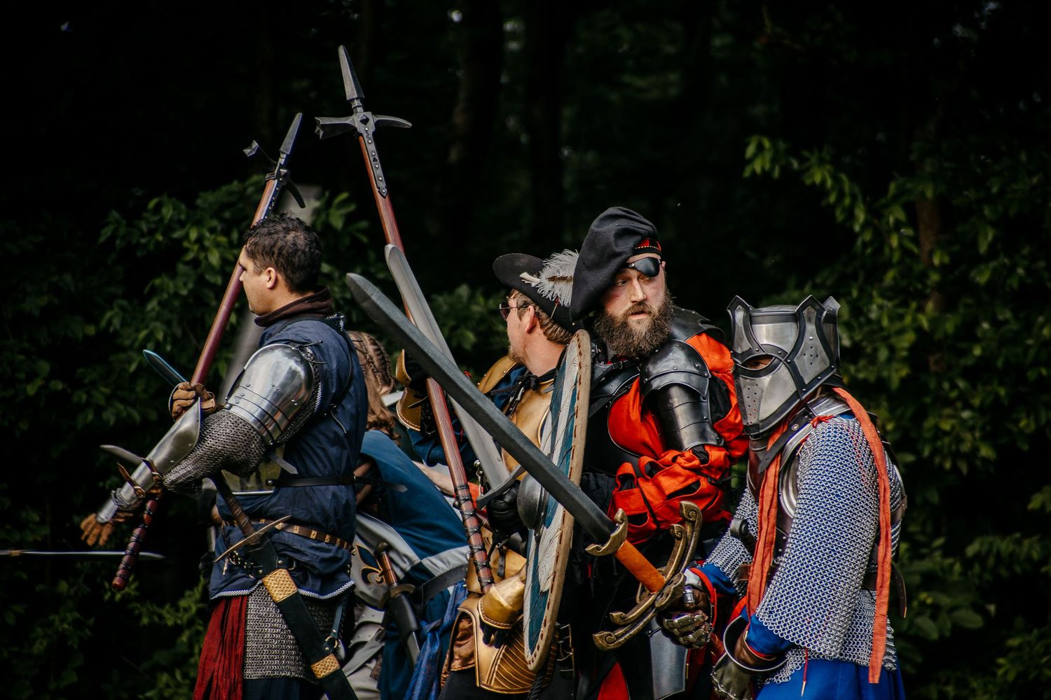 Medieval knights in armor carrying swords and polearms pose dramatically outdoors at a historical reenactment event.
