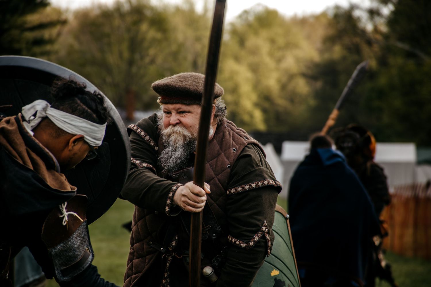 Bearded man in medieval armor wielding a spear during a historical reenactment battle outdoors.