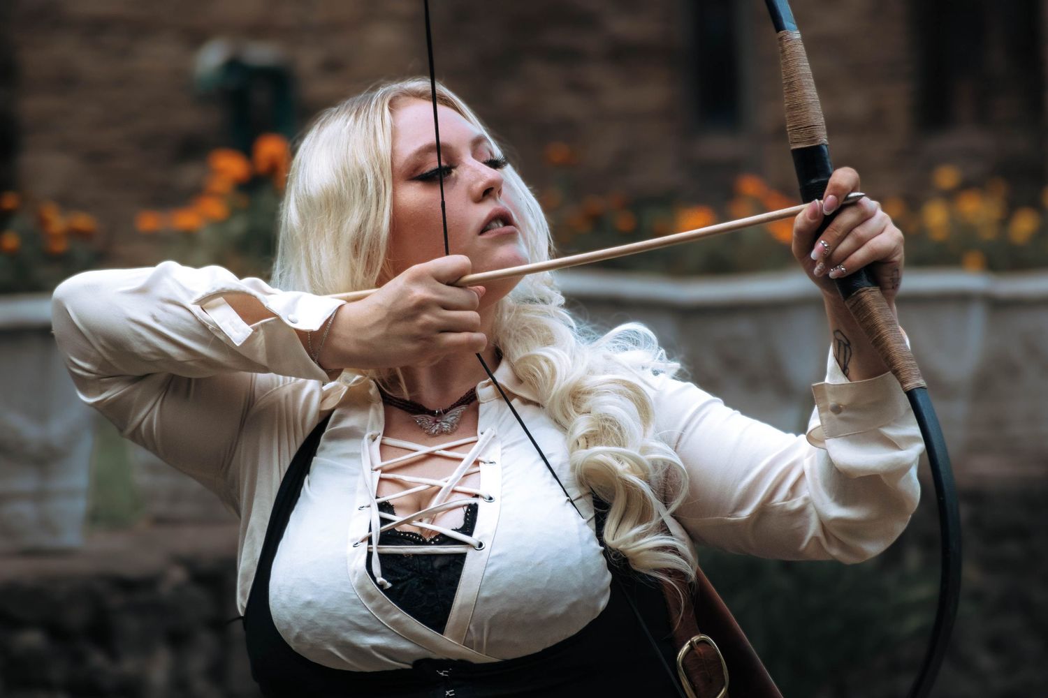 A blonde woman in medieval costume draws a bow and arrow with focused intensity at a Renaissance fair.