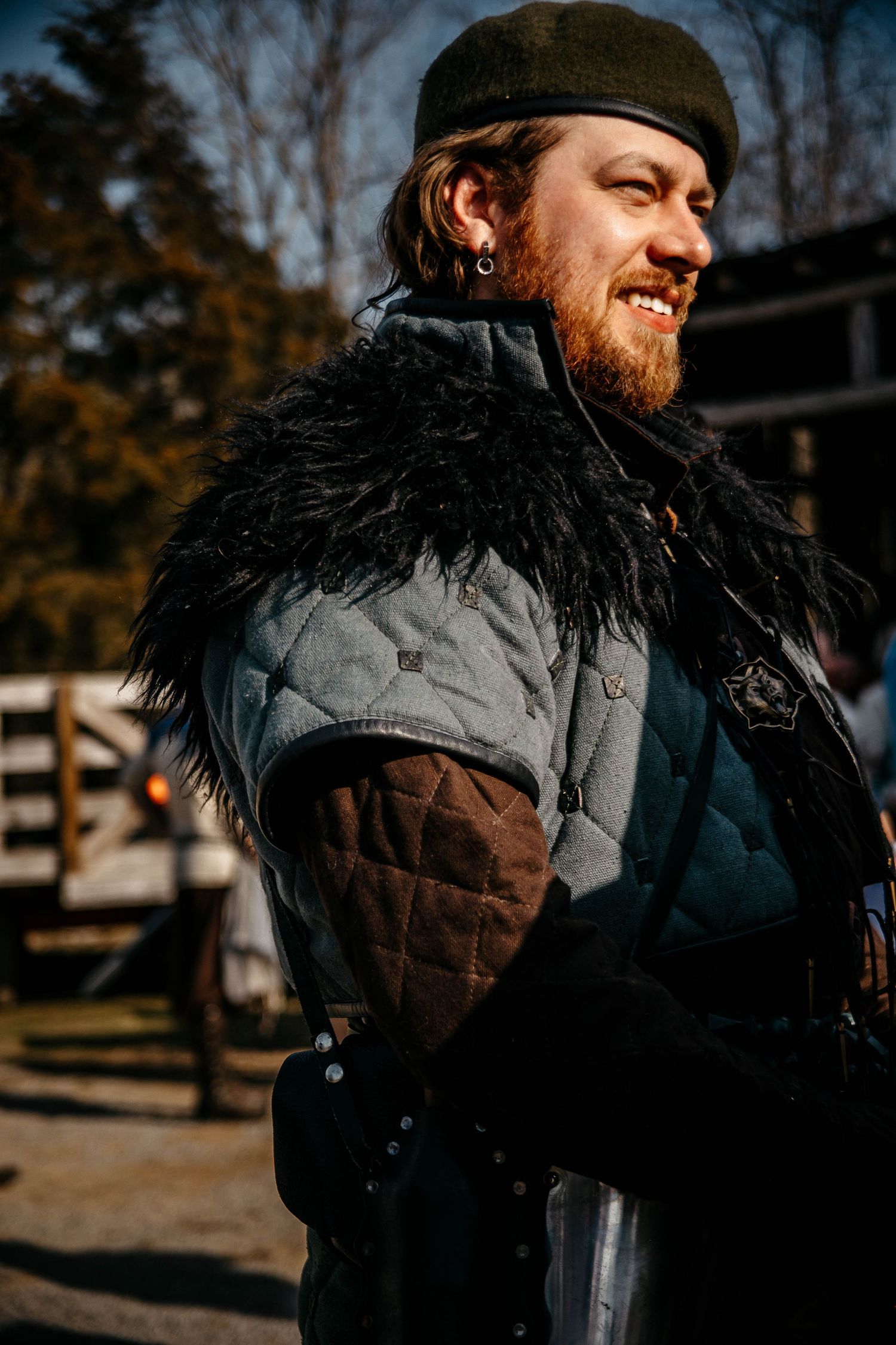 A person wearing a black and brown winter coat with fur trim stands outdoors on an autumn day.