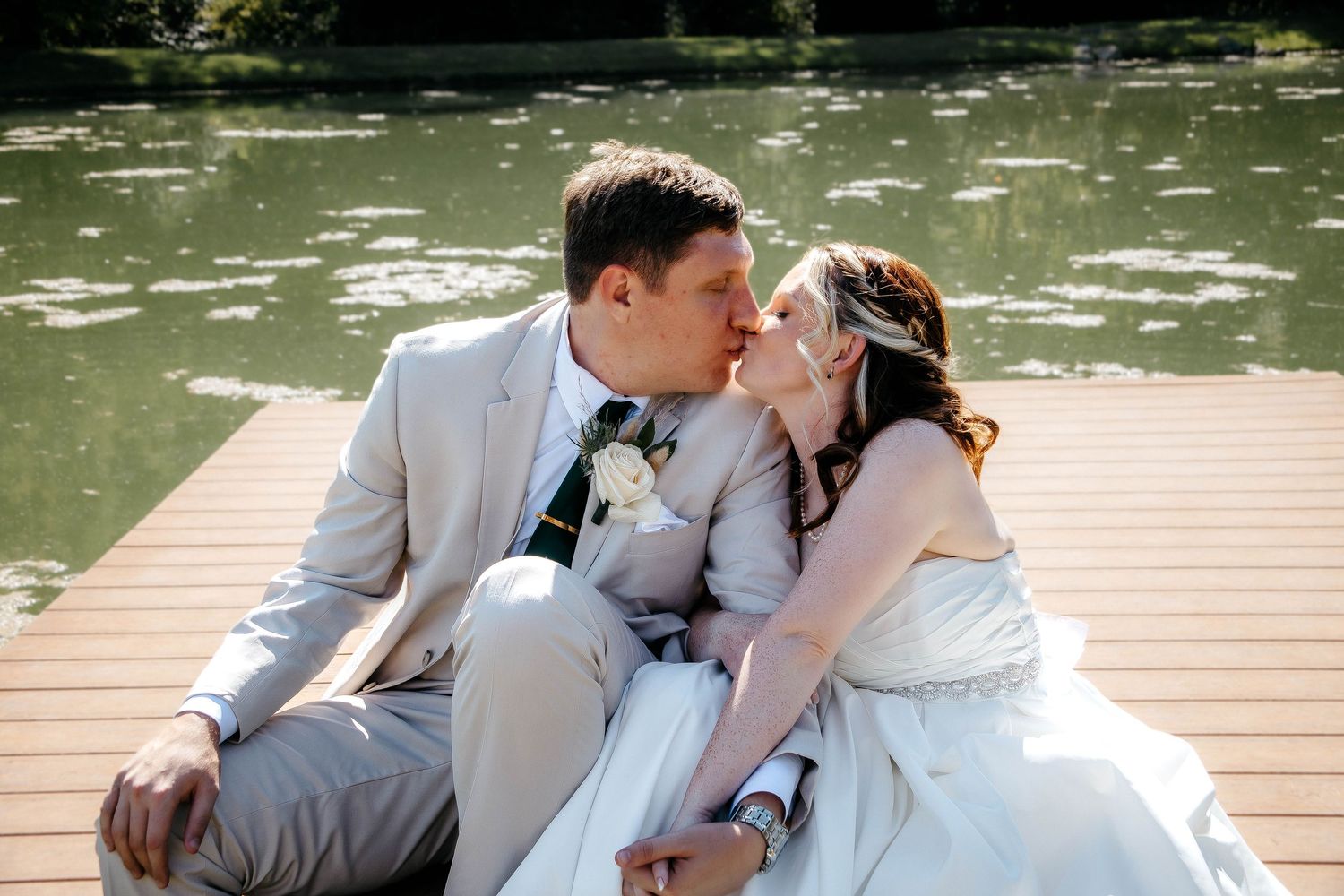Bride and groom sharing a kiss while sitting on a wooden dock by a serene green lake on their wedding day.