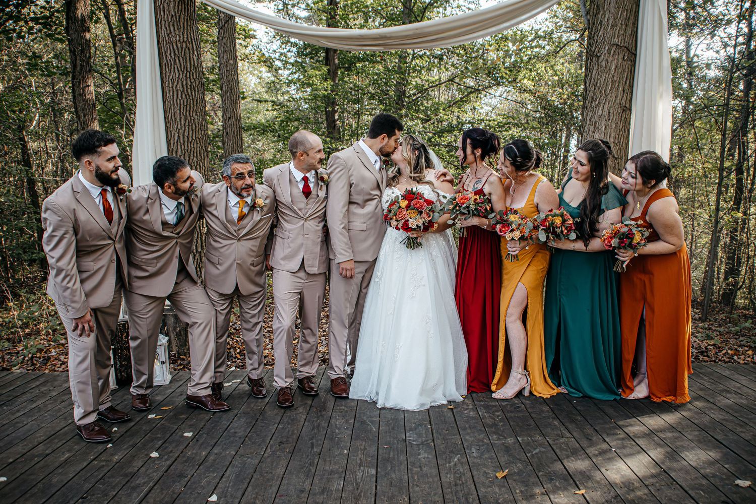 Wedding party in colorful mismatched bridesmaid dresses and tan suits celebrate on a forest deck.