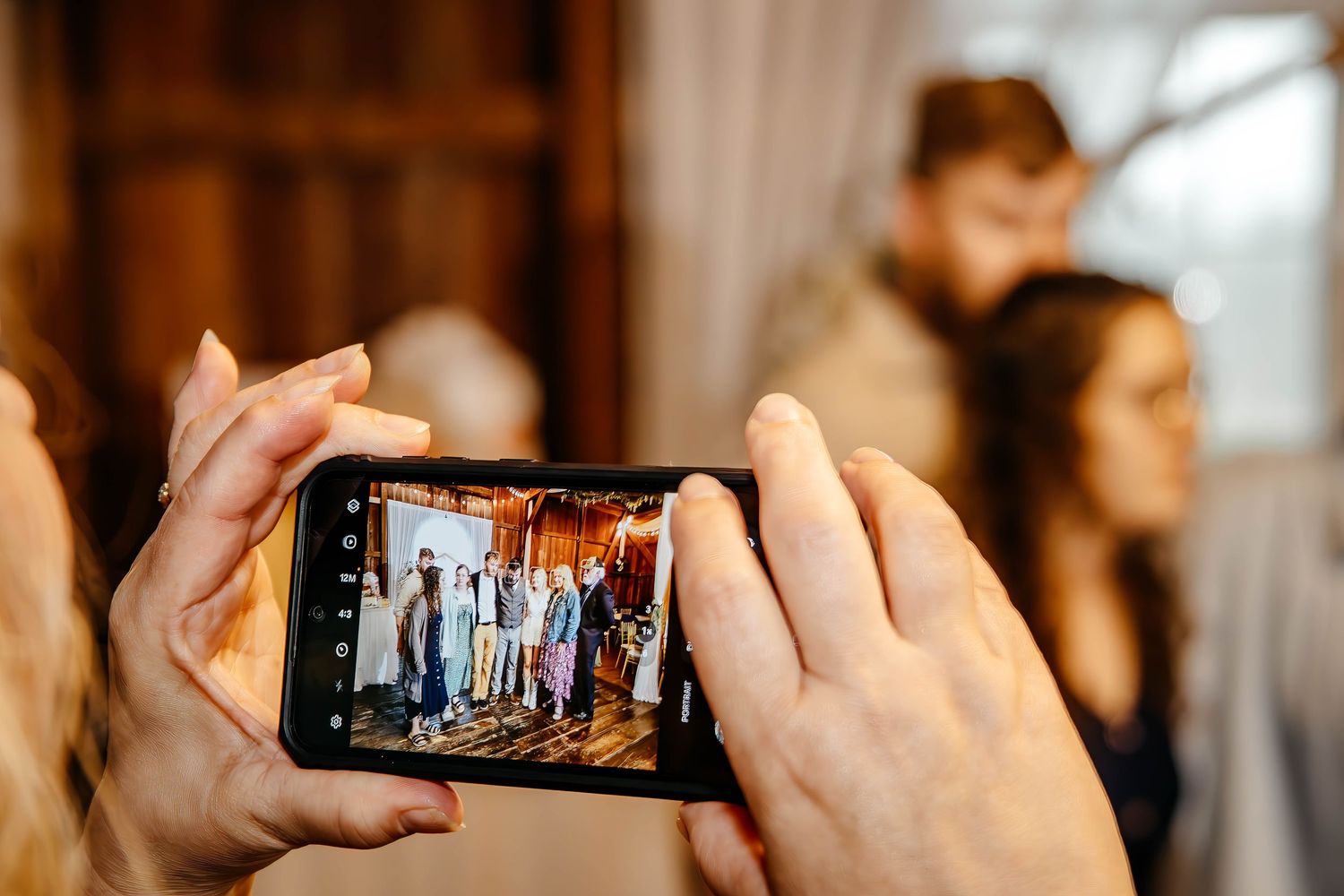 Hands holding a smartphone capturing a group photo at a wedding reception in a rustic venue.
