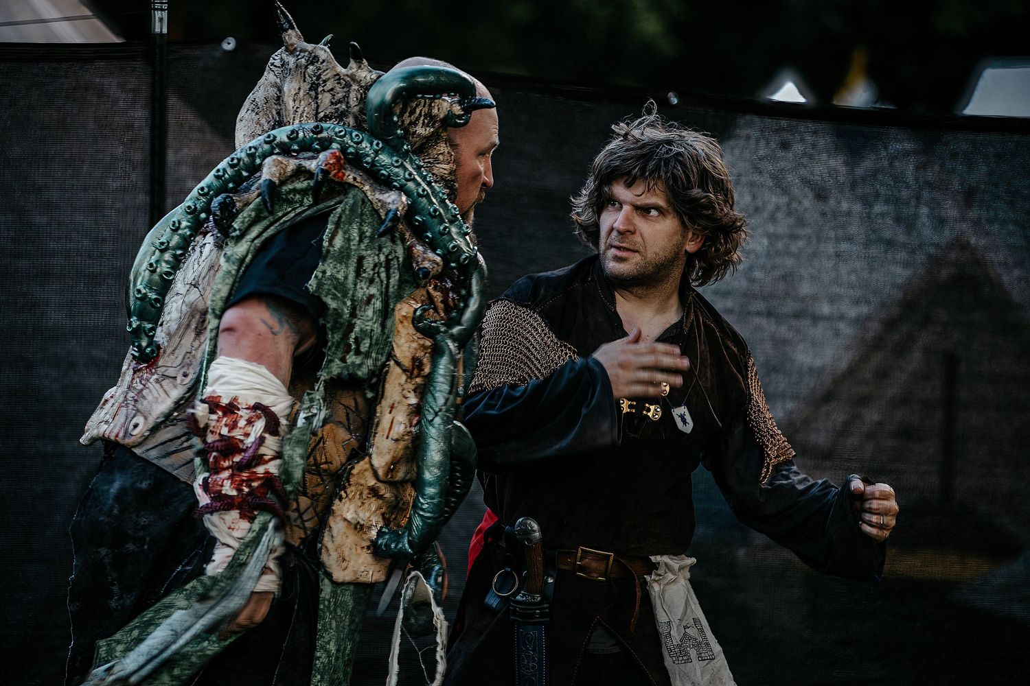 Two actors in medieval fantasy costumes perform a dramatic scene outdoors at a Renaissance fair or theatrical event.