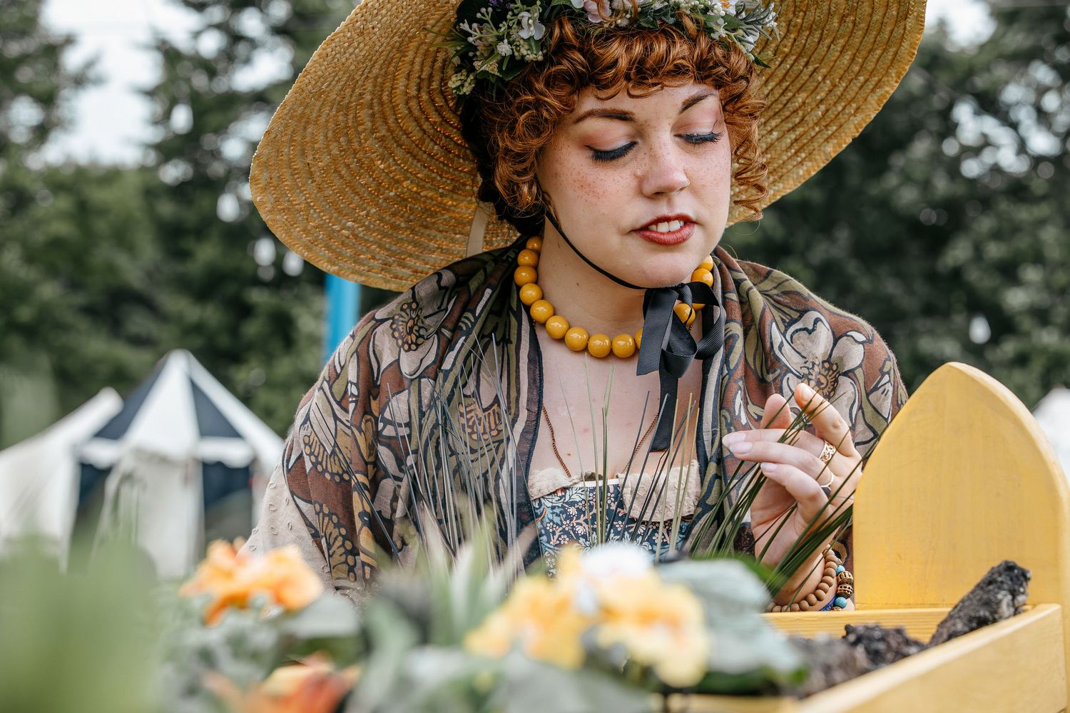 Young woman in straw hat and floral crown with yellow bead necklace and tattoos arranging flowers outdoors.