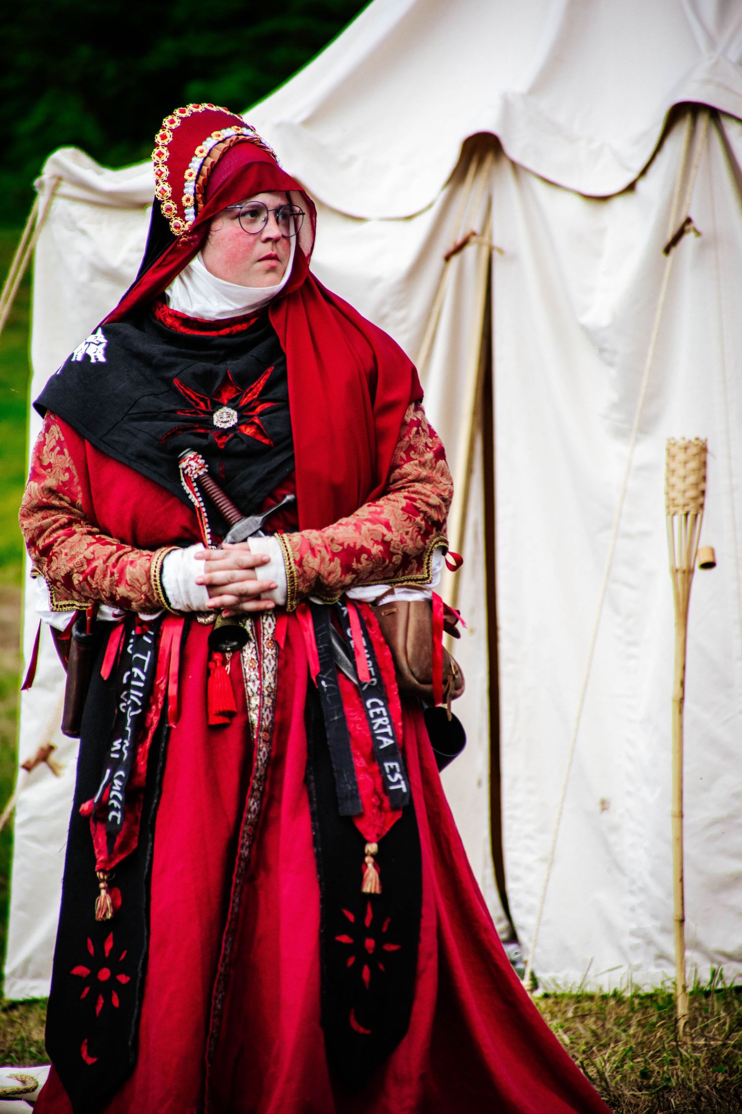 Medieval reenactors in red period costumes with black accents stand near white canvas tents at a historical festival.