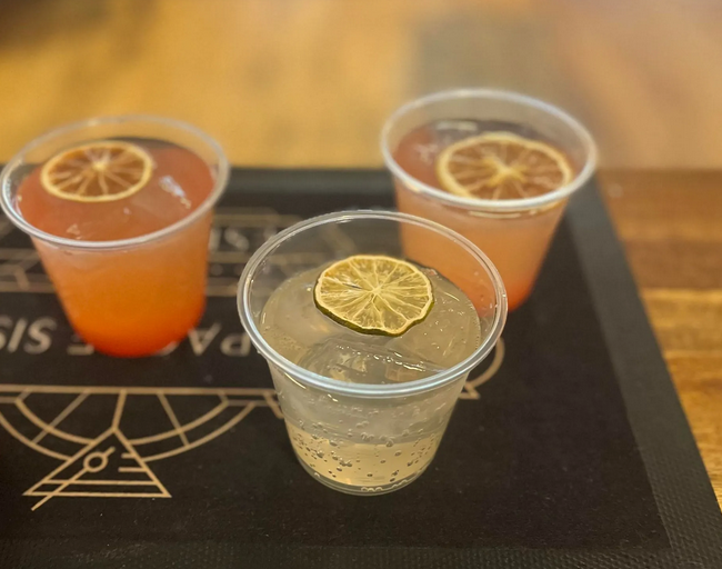A row of colorful craft cocktails garnished with citrus slices sits on a wooden bar top.
