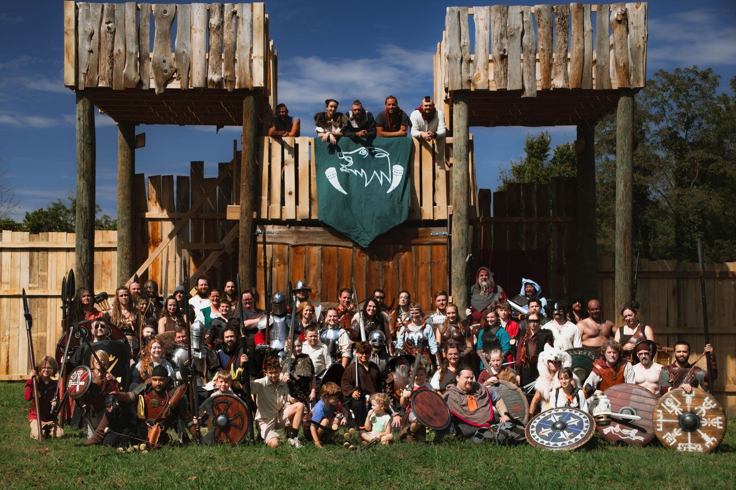 Large group of people gathered outside a rustic wooden structure for a group photo during a sunny day.