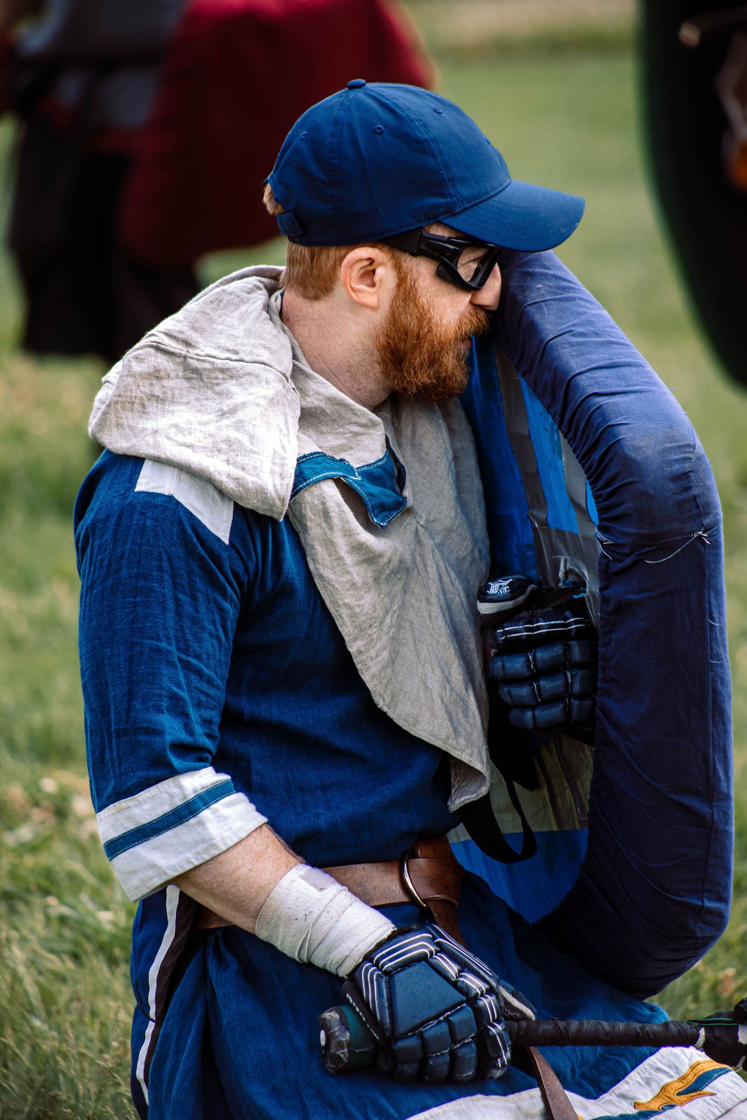 A person wearing a blue baseball cap and vest sits on grass while looking at a book or magazine.