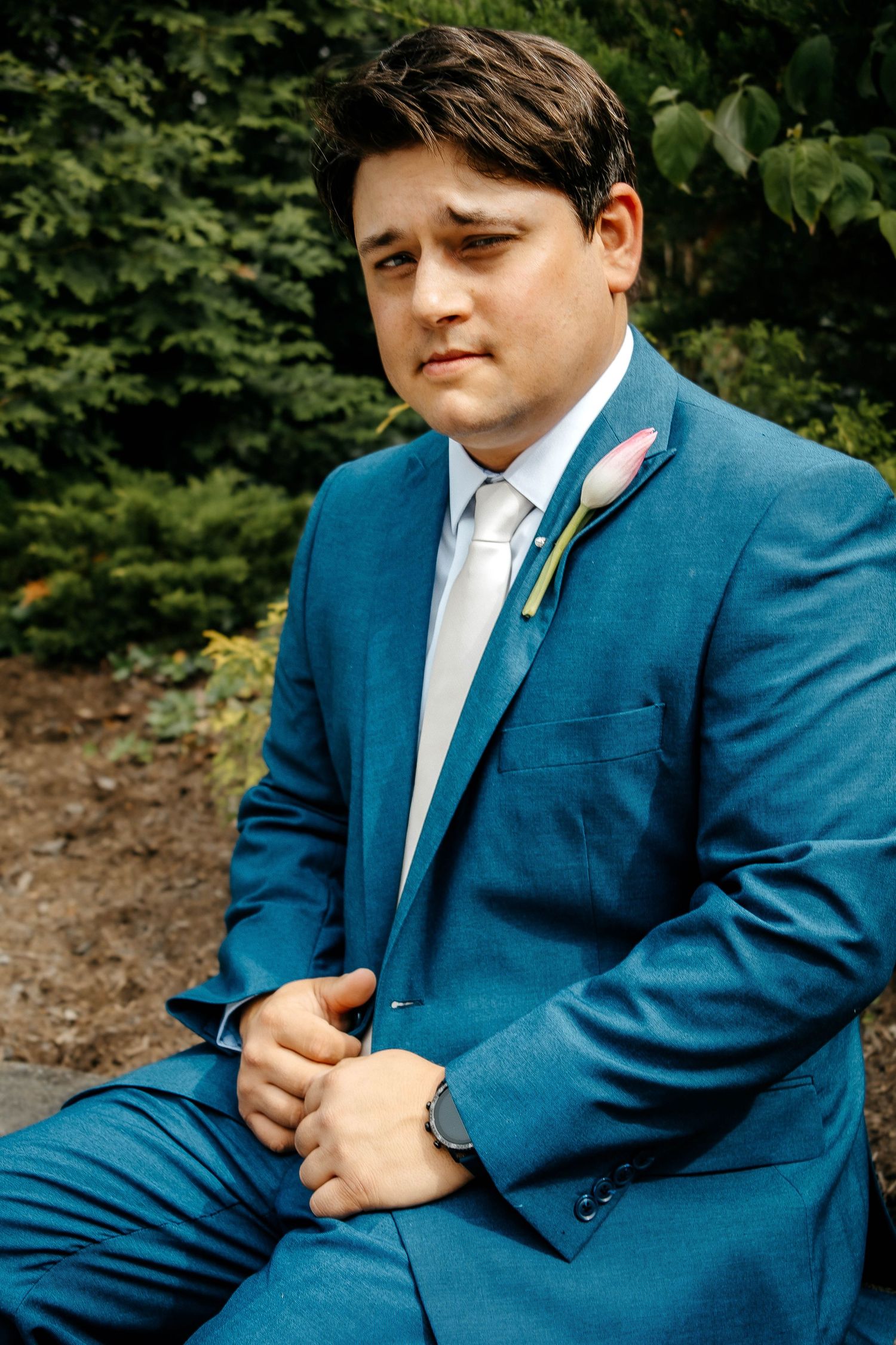A person in a teal blue suit and white dress shirt adjusts their tie while sitting outdoors.