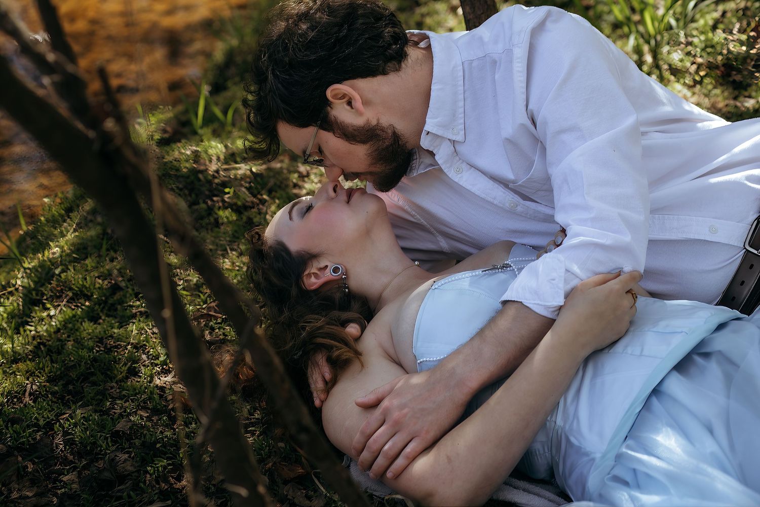 A romantic couple shares a tender kiss outdoors, with the woman lying on grass and the man leaning over her.
