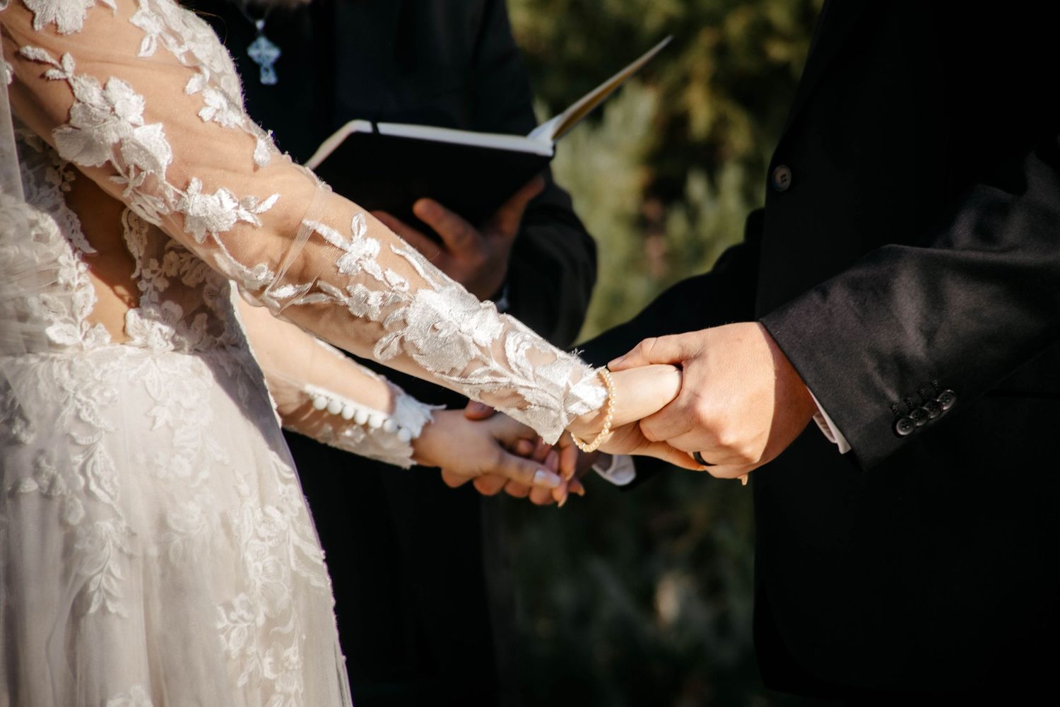 Close-up of hands exchanging wedding rings during outdoor ceremony with lace dress detail and officiant holding book.