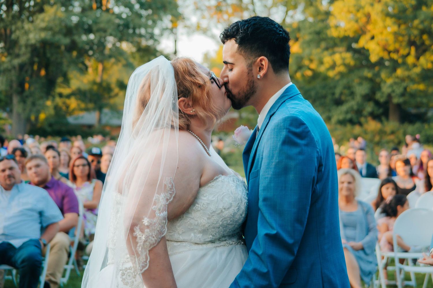 A couple shares a romantic moment during their outdoor wedding ceremony with autumn foliage in the background.