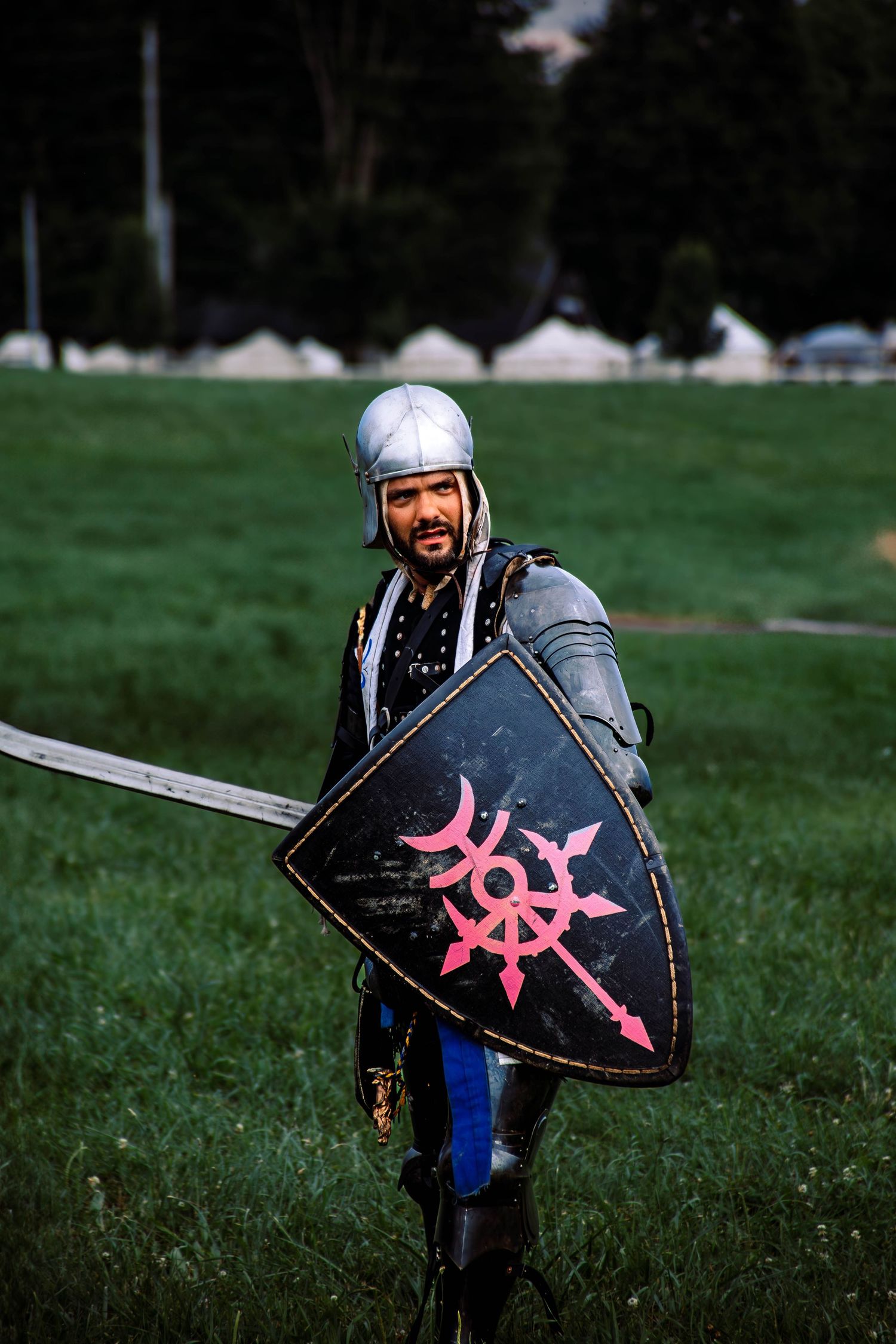 A medieval cosplayer in armor holds a black shield with pink markings while standing in a grassy field.