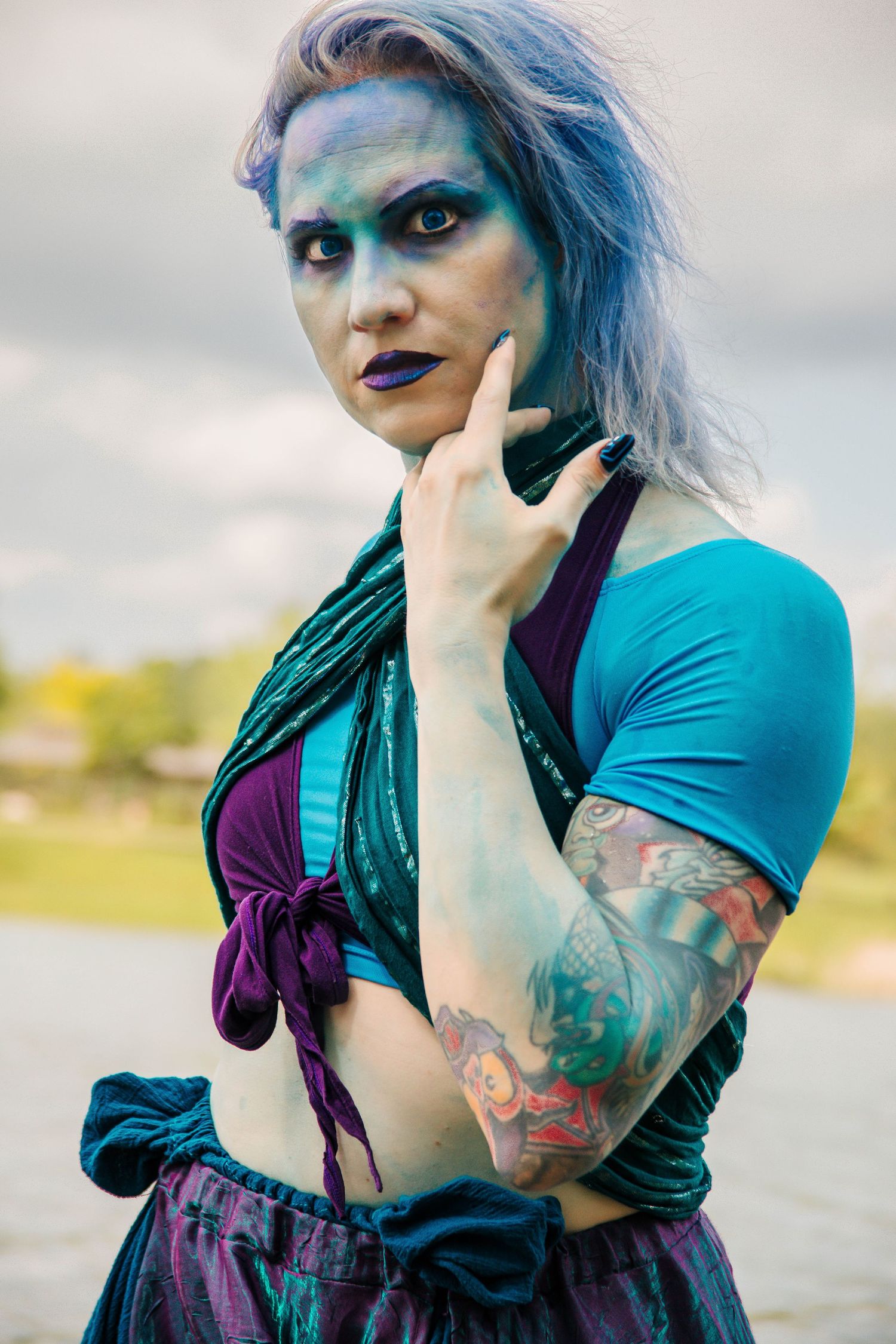 Woman with blue hair and body paint, teal crop top, tattoos, and purple lipstick poses outdoors under cloudy sky.