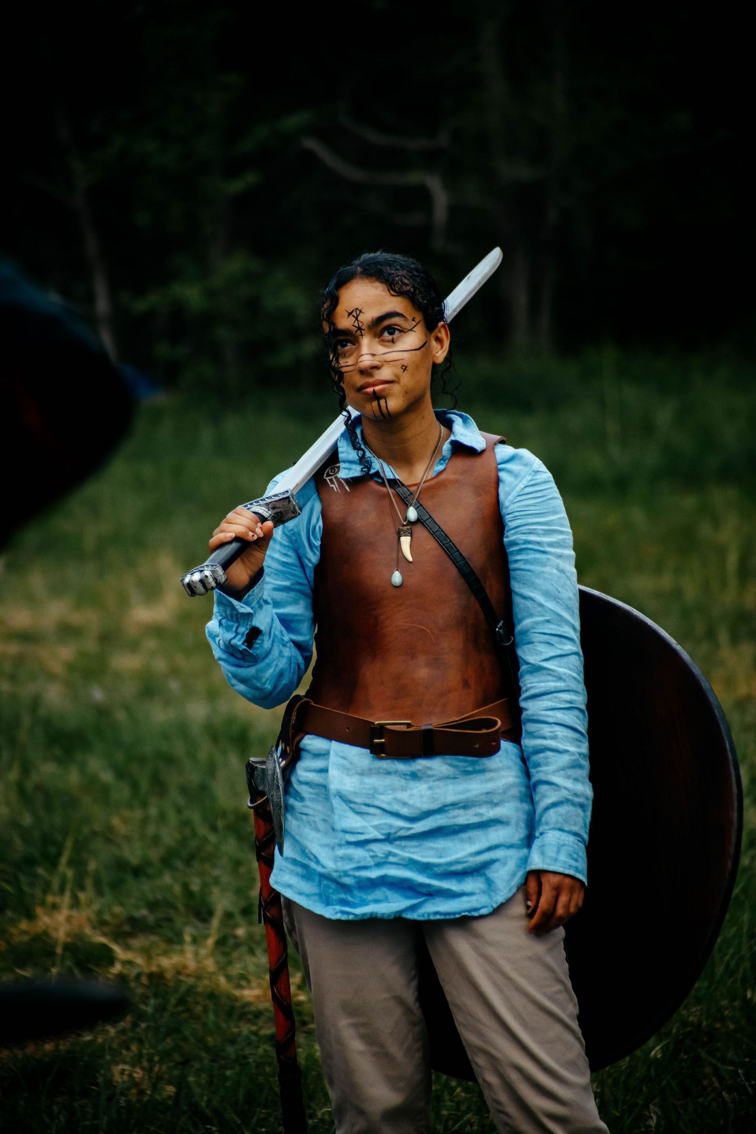 A person in a light blue shirt and brown vest holds a sword while practicing archery outdoors.