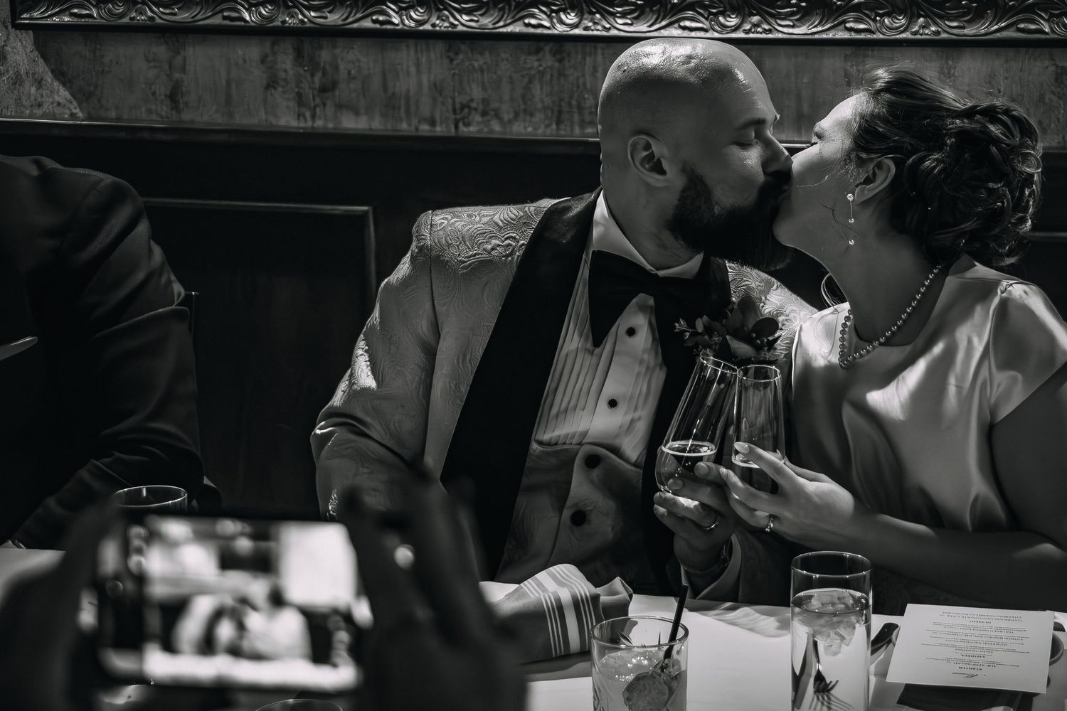 Black and white wedding photo of couple sharing a kiss while holding champagne glasses at reception dinner table.