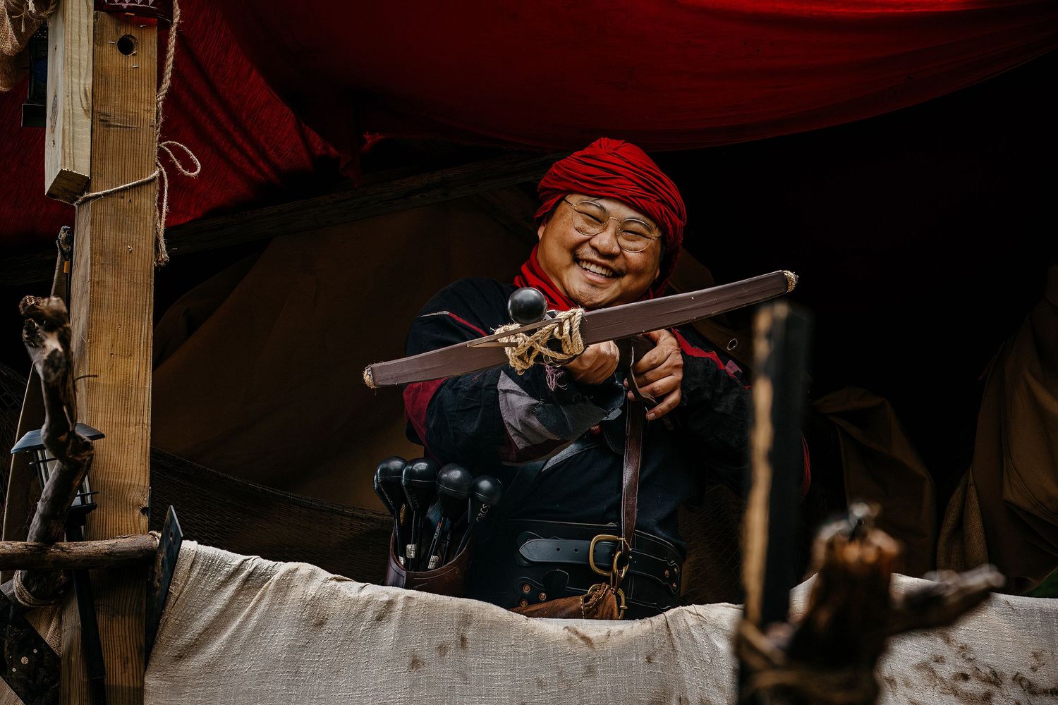 Elderly Asian woman in red headscarf smiling while weaving traditional textile on a wooden loom outdoors.