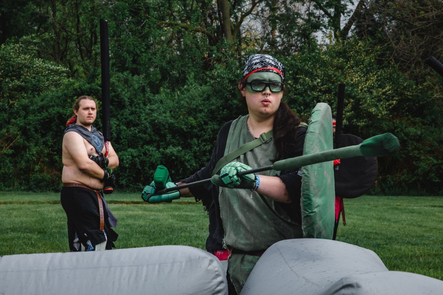 Two people playing outdoor inflatable combat game on grass, one holding green foam weapon near gray barrier.