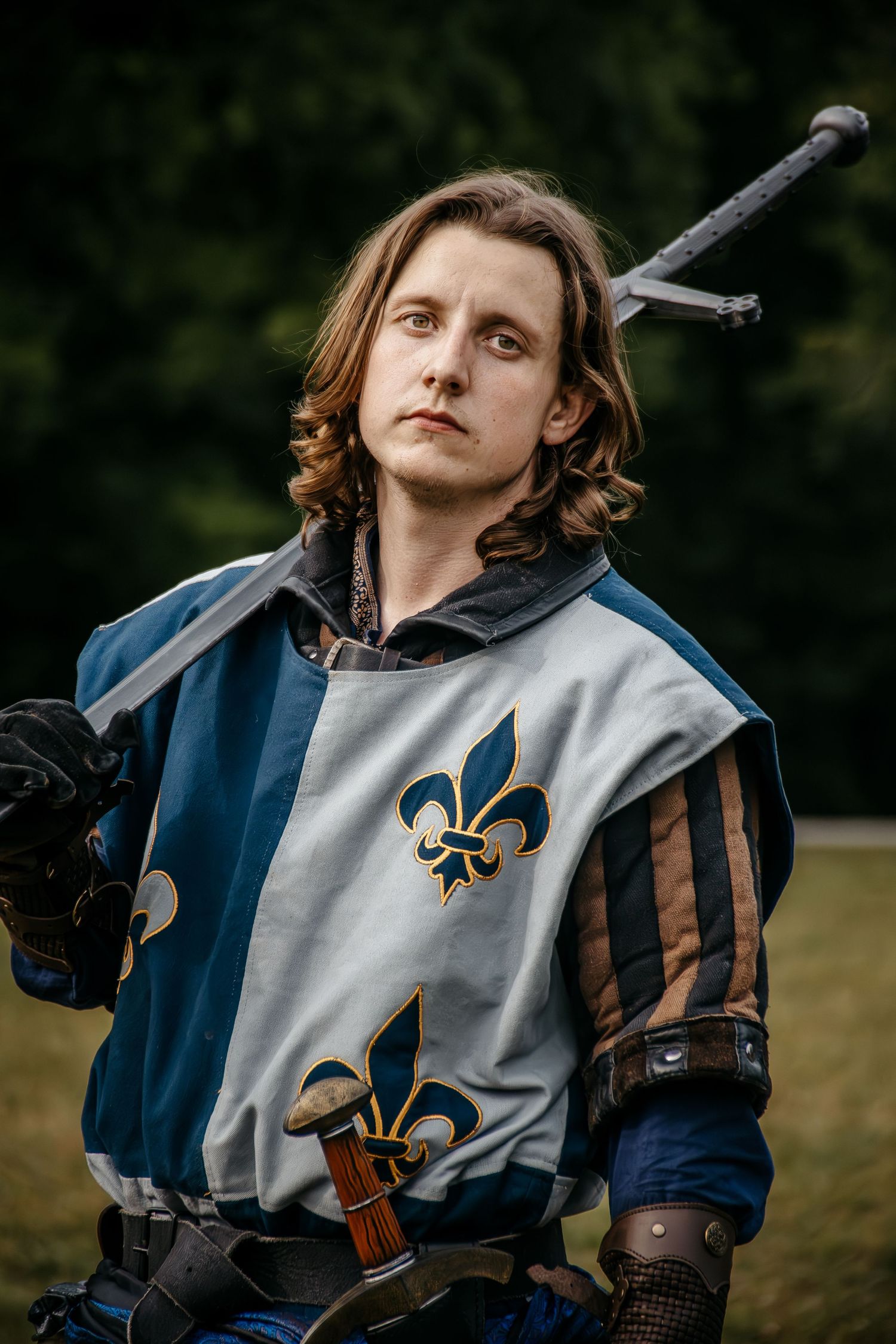 Young man in medieval knight costume with fleur-de-lis tabard and sword, posing outdoors.