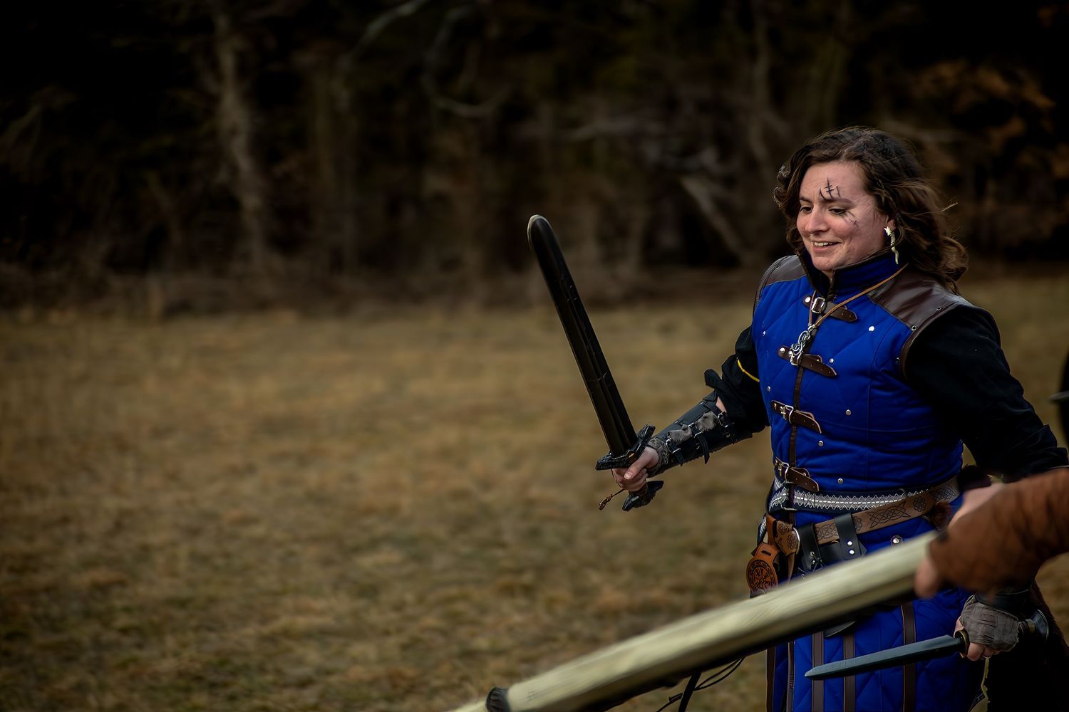 Woman in blue medieval armor costume wielding a black sword outdoors at a LARP or cosplay event.
