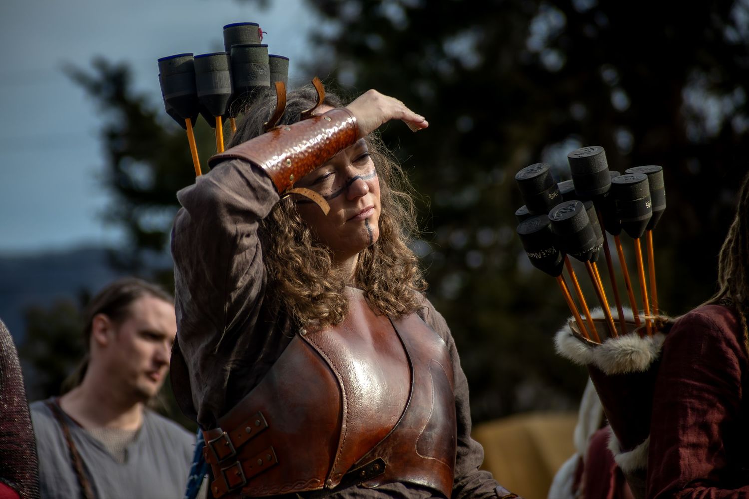 Medieval warrior woman in leather armor with arrows, shielding eyes at outdoor Renaissance faire event.