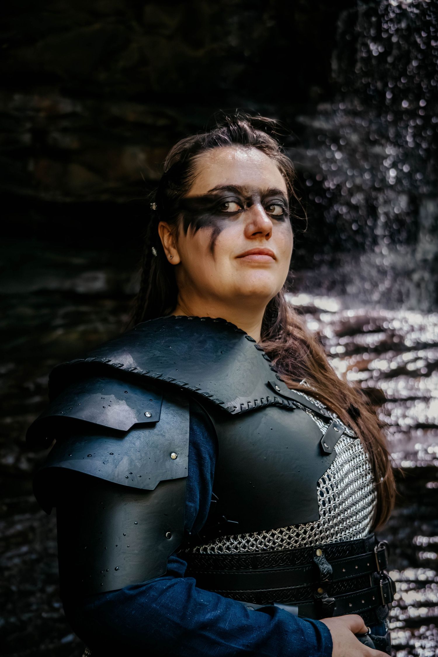 A person wearing medieval-style black leather and chainmail armor poses against a dark stone background.