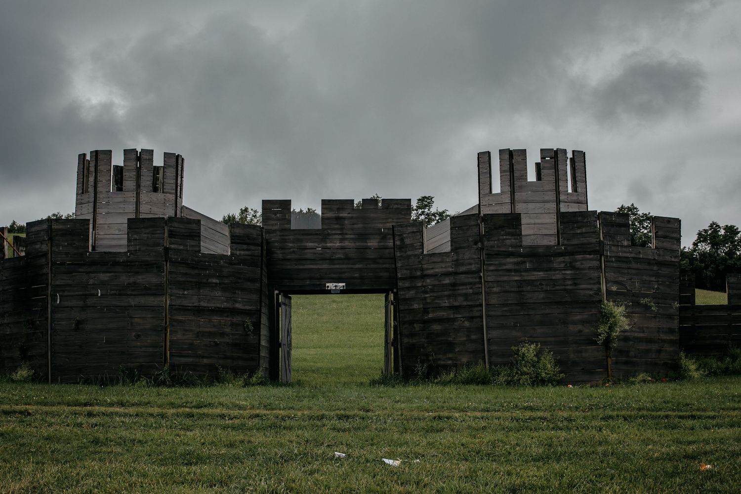 A dark wooden medieval castle structure with battlements stands on a grassy field under a cloudy sky.