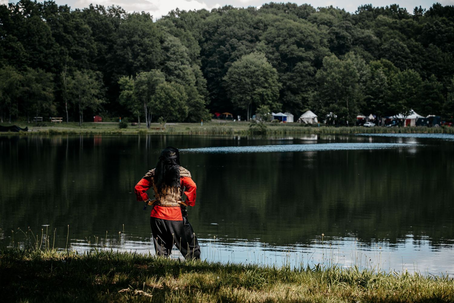 Person in orange hoodie standing by a calm reflective lake surrounded by lush green forest trees.