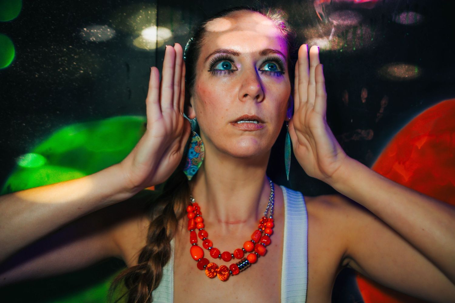 Woman with colorful makeup, orange beaded necklace, and blue earrings posing against a vibrant neon background.