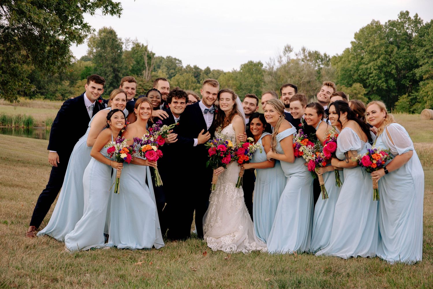 A wedding party in light blue bridesmaid dresses and red bouquets poses together outdoors in a natural setting.