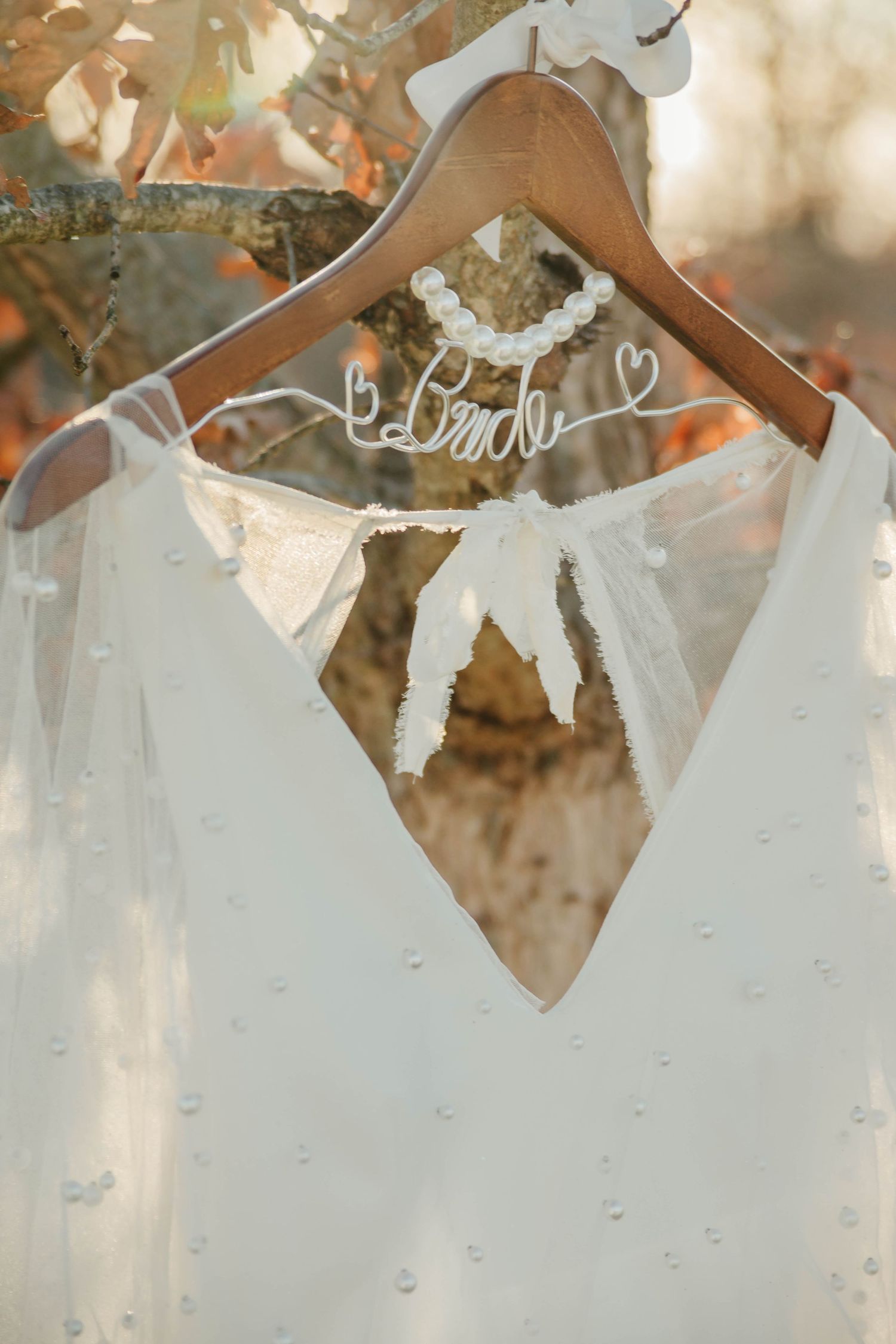 A white wedding dress with a V-neckline hangs on a personalized wooden hanger with hearts against a soft autumn background.