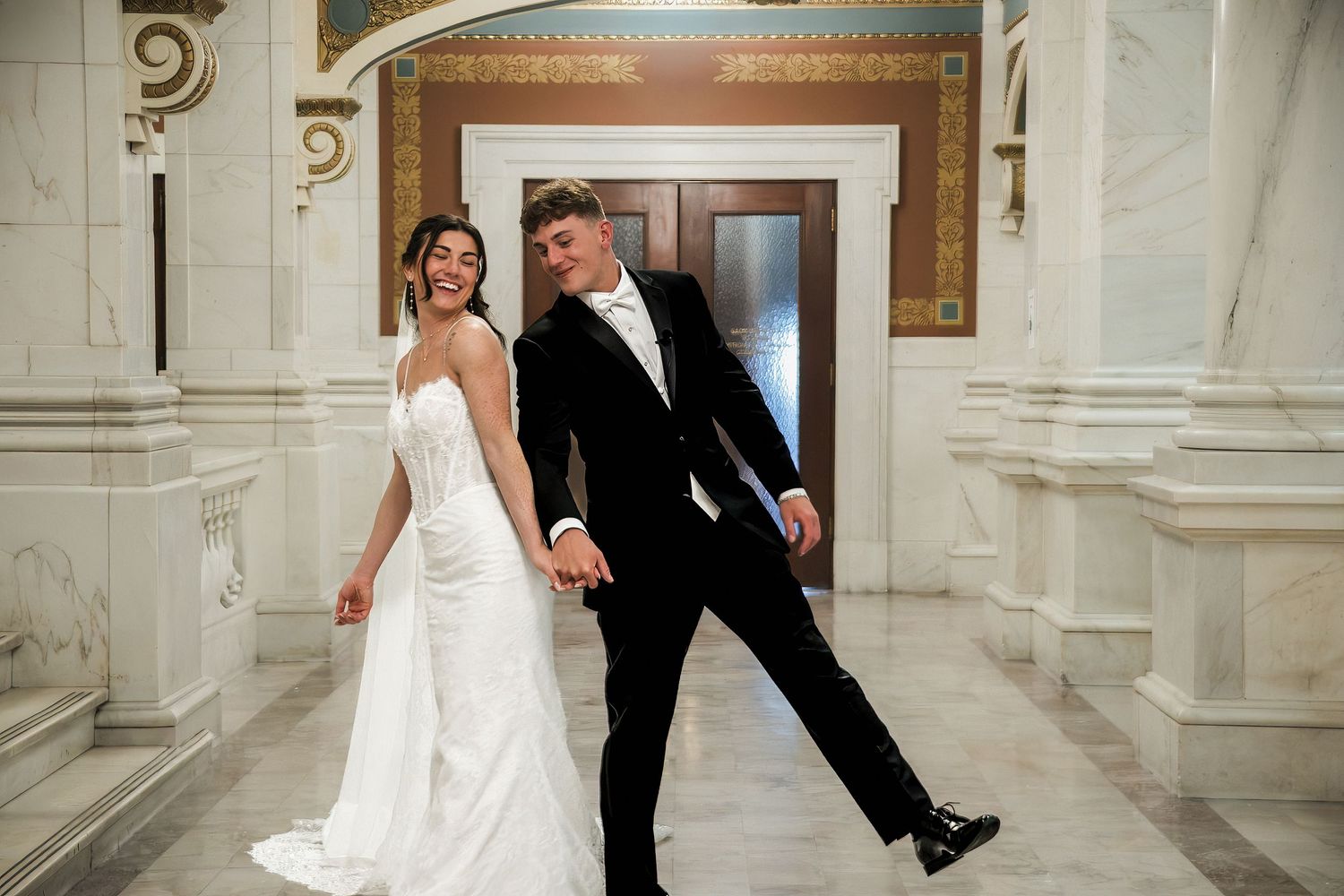 Bride in white gown and groom in black tuxedo hold hands and pose playfully in an elegant marble hall.