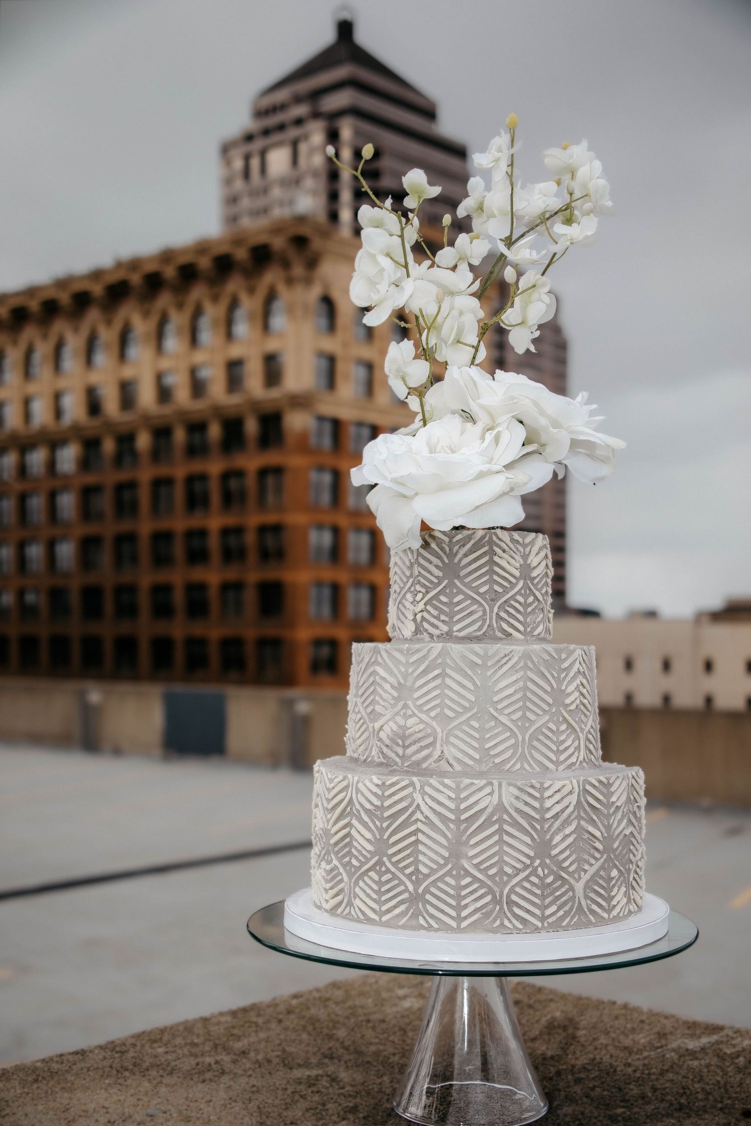 Three-tier geometric patterned wedding cake with white floral topper displayed on rooftop with urban building backdrop.