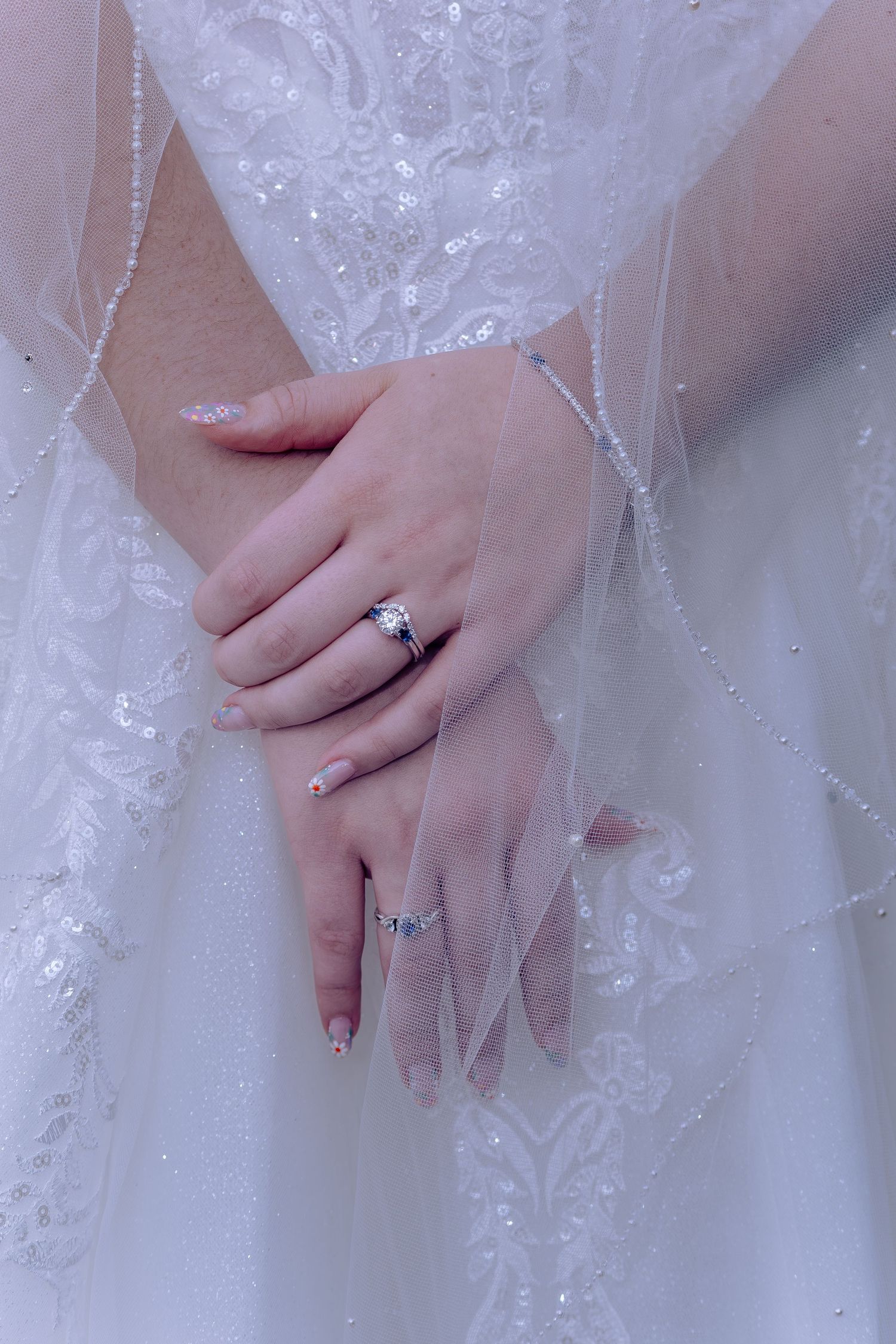 Close-up view of hands wearing an elegant engagement ring against white lace wedding dress fabric.