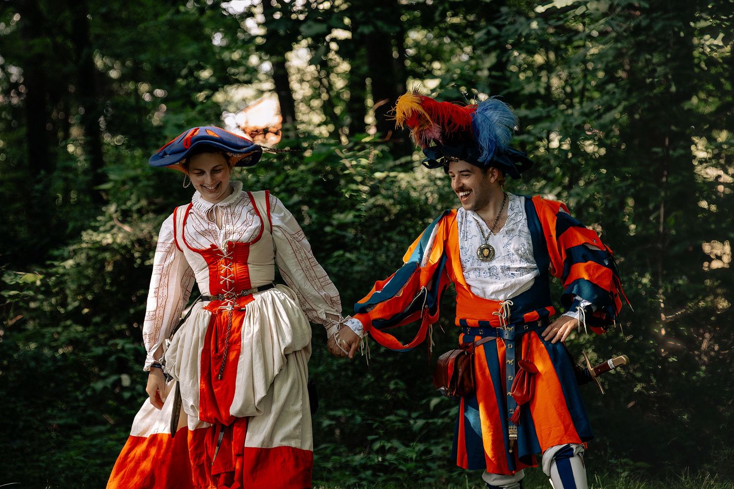 Two people in colorful Renaissance costumes holding hands and walking through a lush green forest.