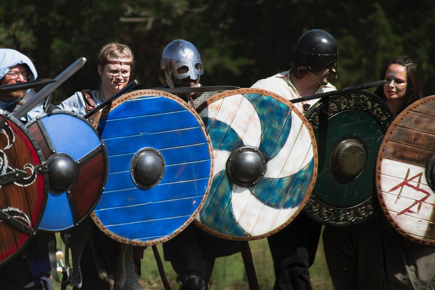 A line of medieval warriors with blue and white shields stand in formation during a historical reenactment event.