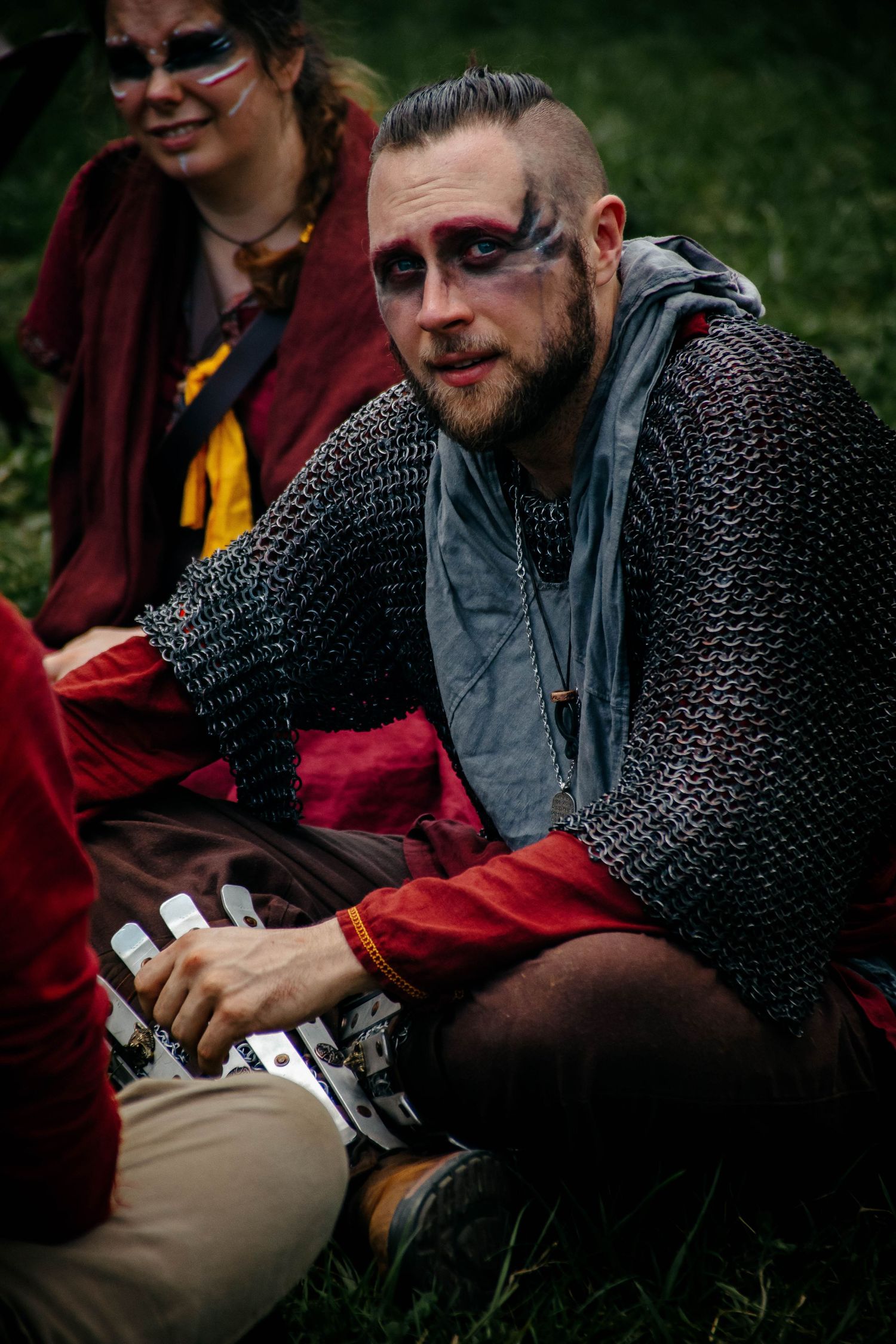 Medieval reenactor in chainmail armor and modern clothing relaxes on grass during outdoor event.