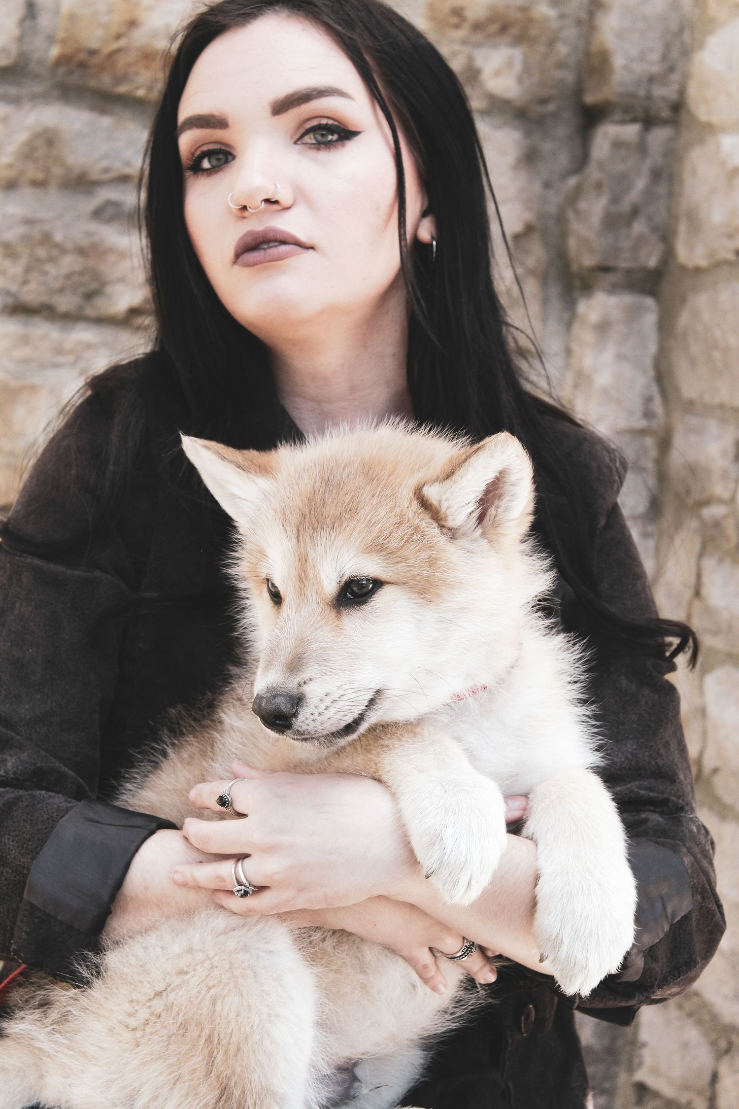 Person in dark clothing cuddles with fluffy cream-colored fox against stone wall.