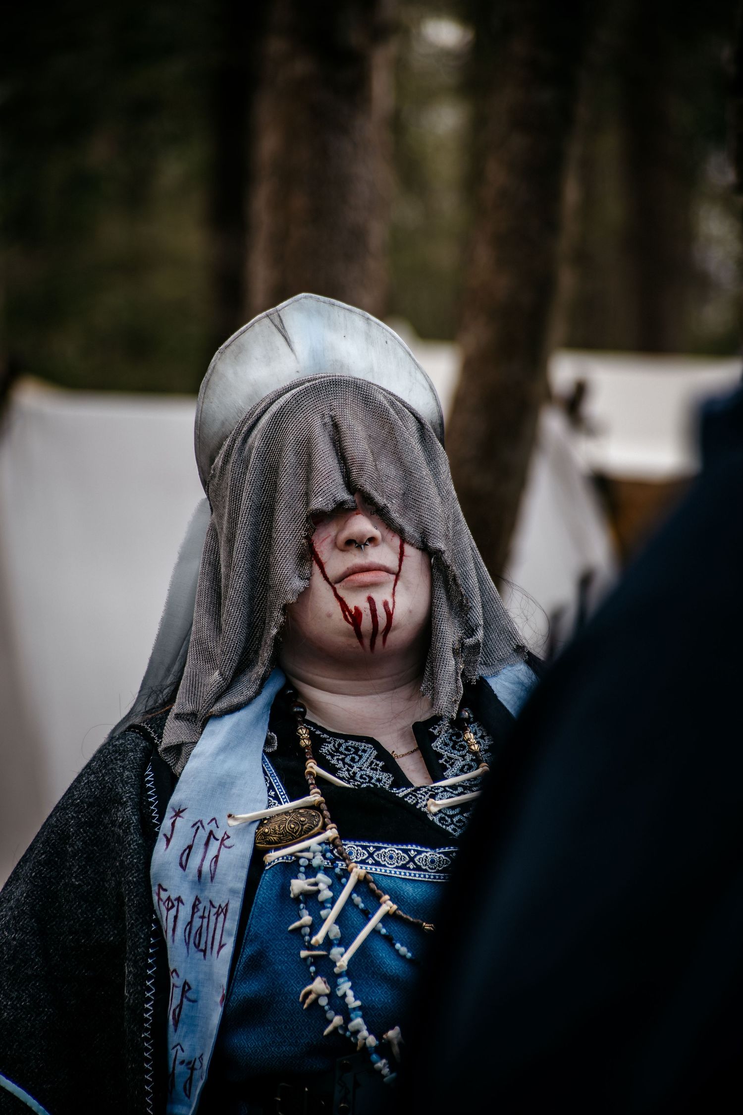 Person in medieval costume with hood and fake blood face makeup at outdoor LARP or Renaissance fair event.