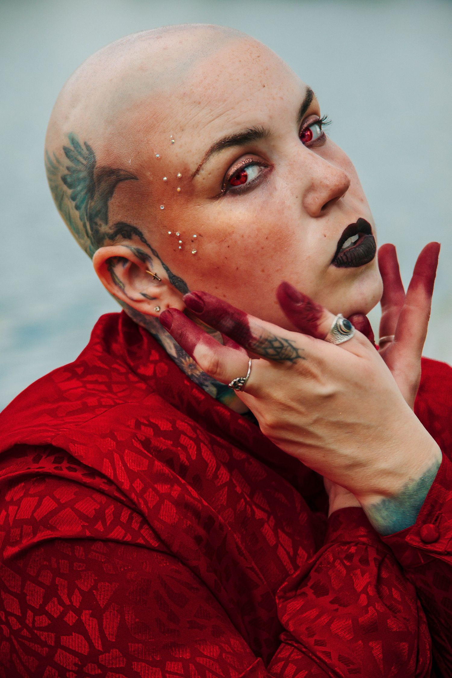 Bald woman with red contact lenses, dark lipstick, tattoos, and red-painted hands poses in a red jacket.