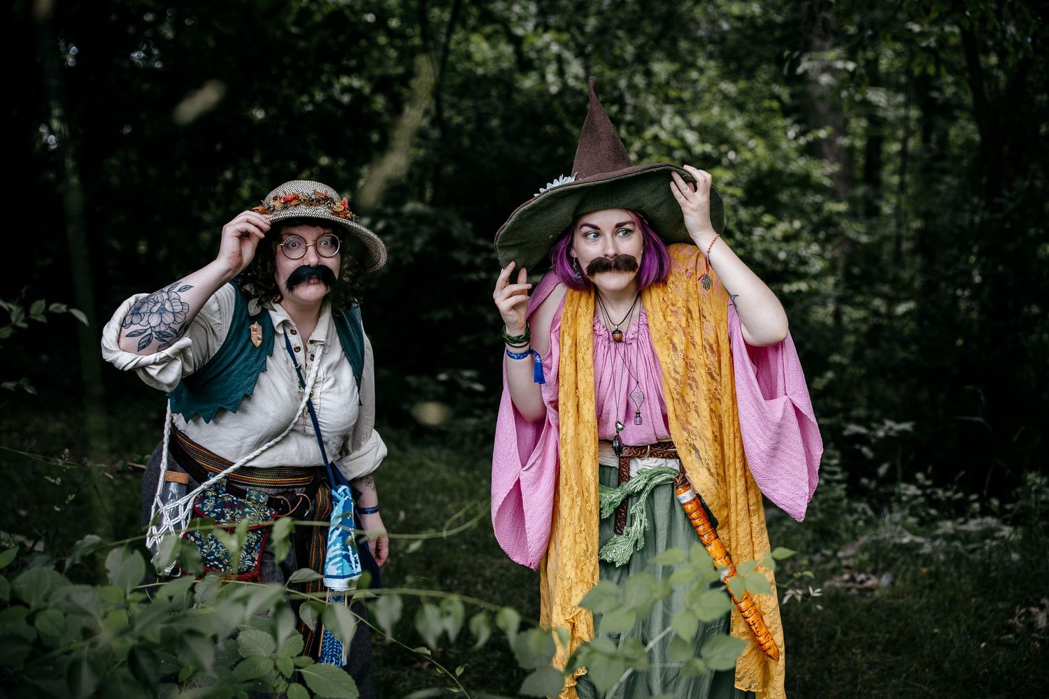 Two people in wizard and pirate costumes with fake mustaches posing in a forest setting.