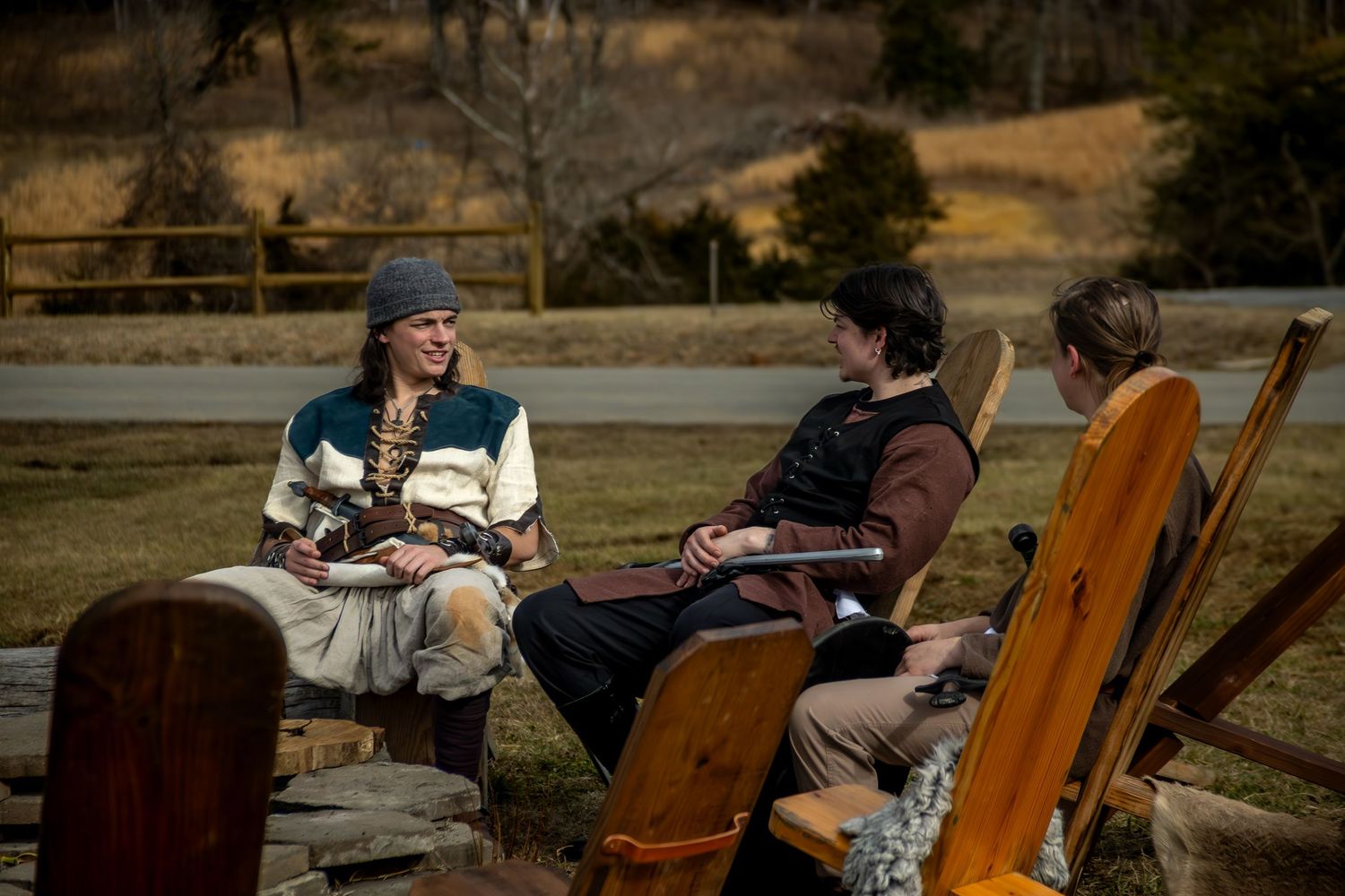 Musicians perform outdoors on stringed instruments while seated in wooden chairs at a rustic outdoor venue.