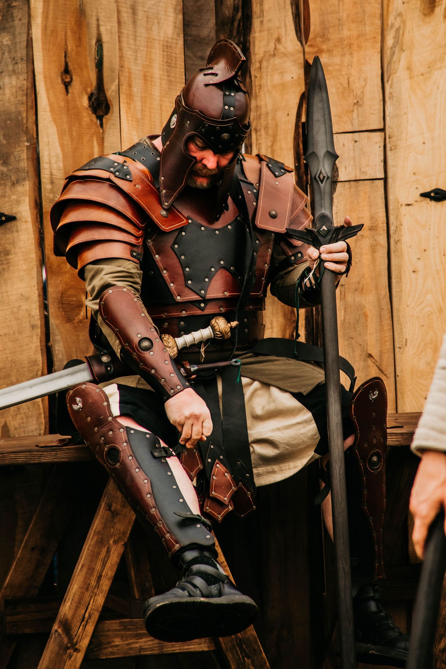 Medieval warriors in leather armor and studded gauntlets prepare their weapons against wooden wall backdrop.